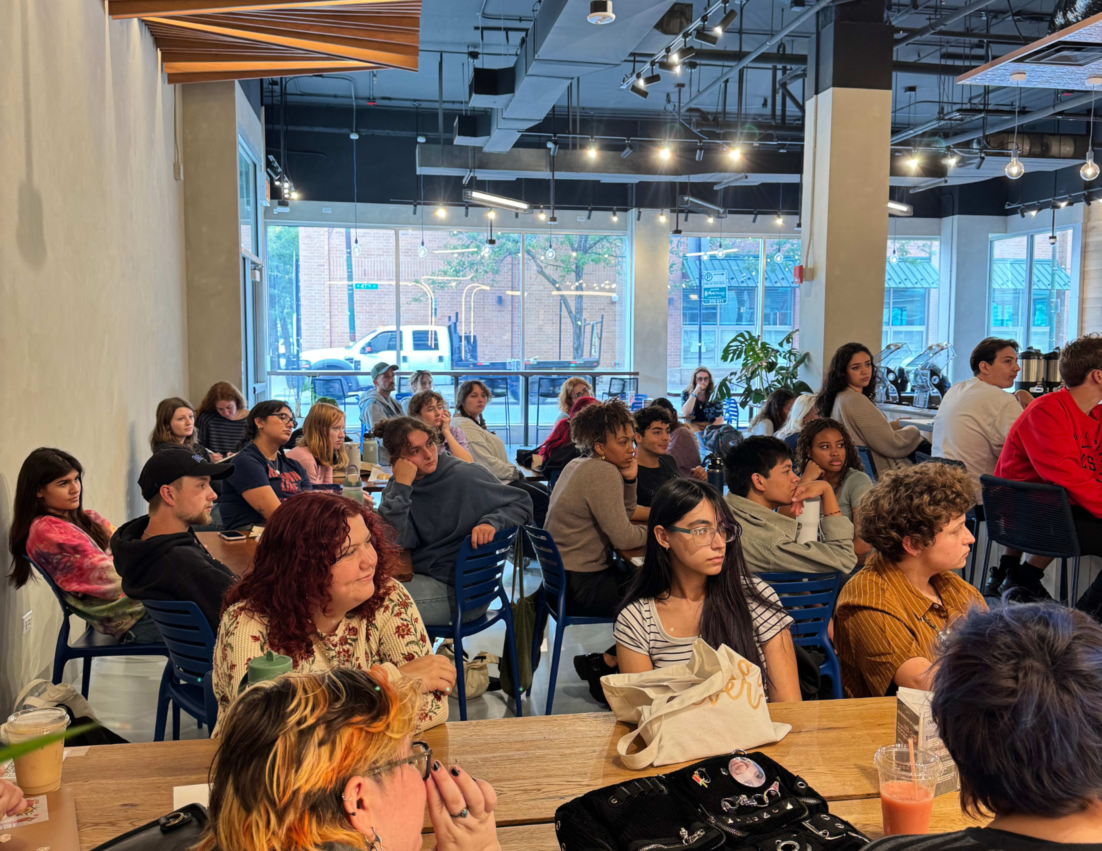 A diverse group of people is seated in a café, attentively facing the front where a presentation appears to be taking place.