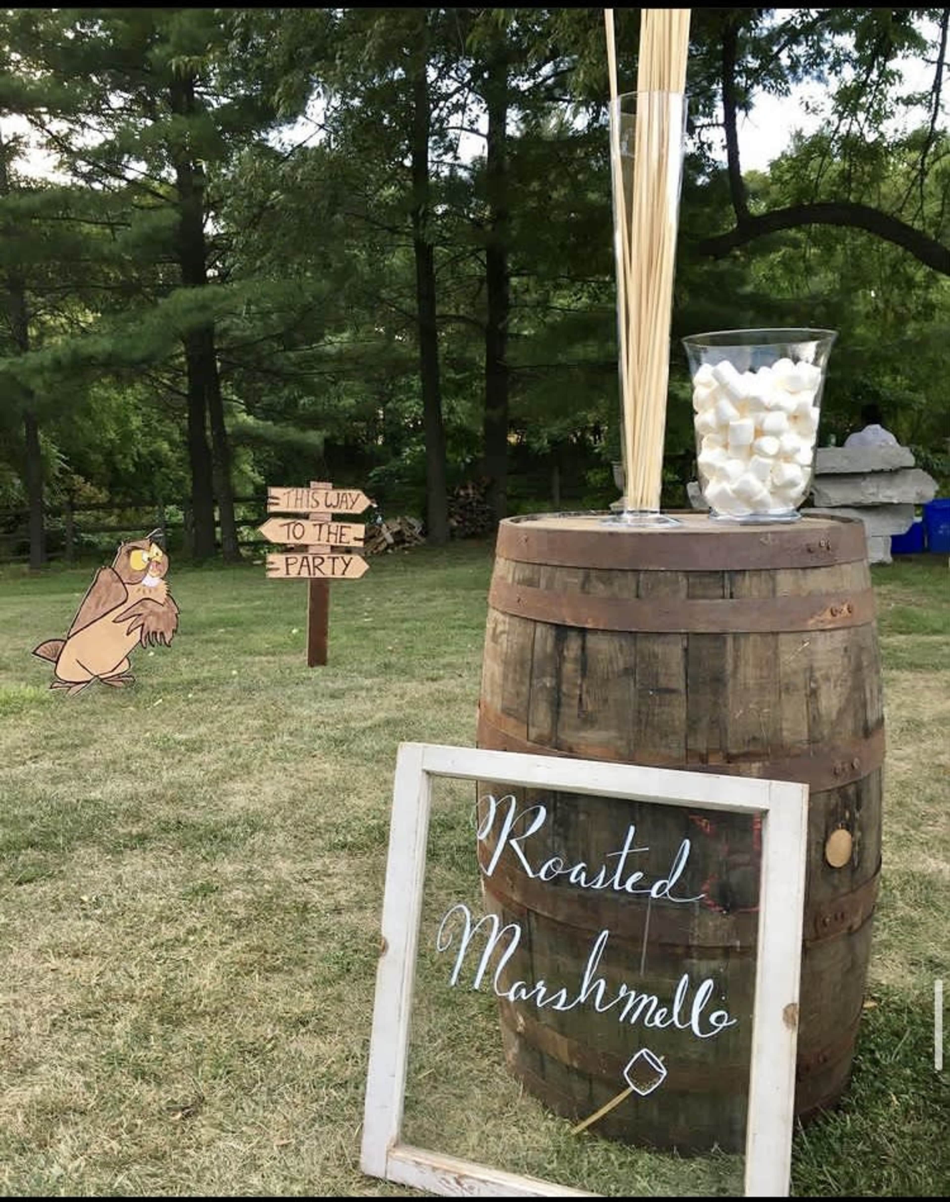 A wooden barrel displays a sign reading "Roasted Marshmelloe" alongside a glass container filled with marshmallows, while a whimsical sign points guests towards the party.
