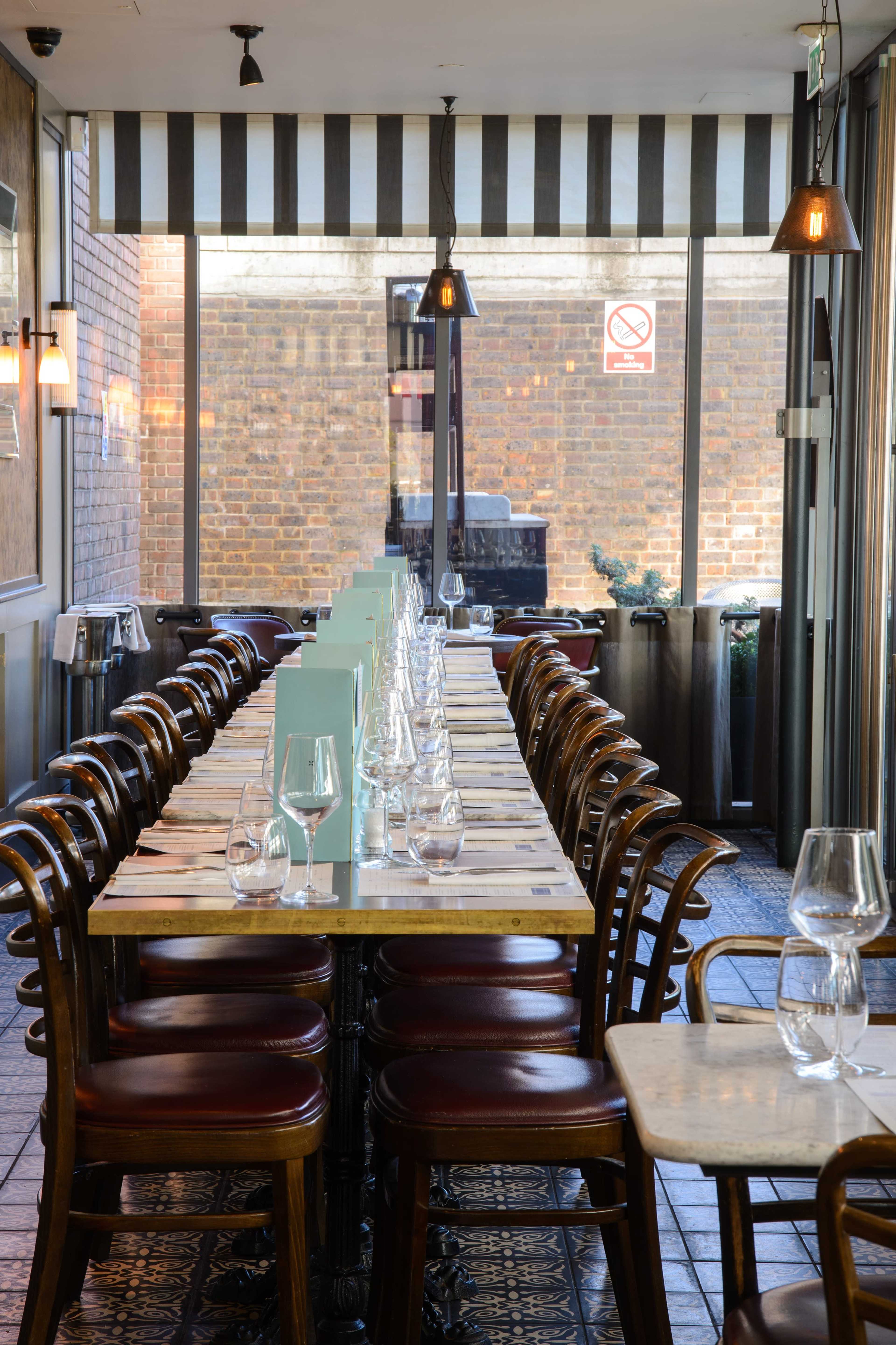 A long, set dining table with empty wine glasses is arranged in a restaurant with large windows and a view of a brick wall outside.