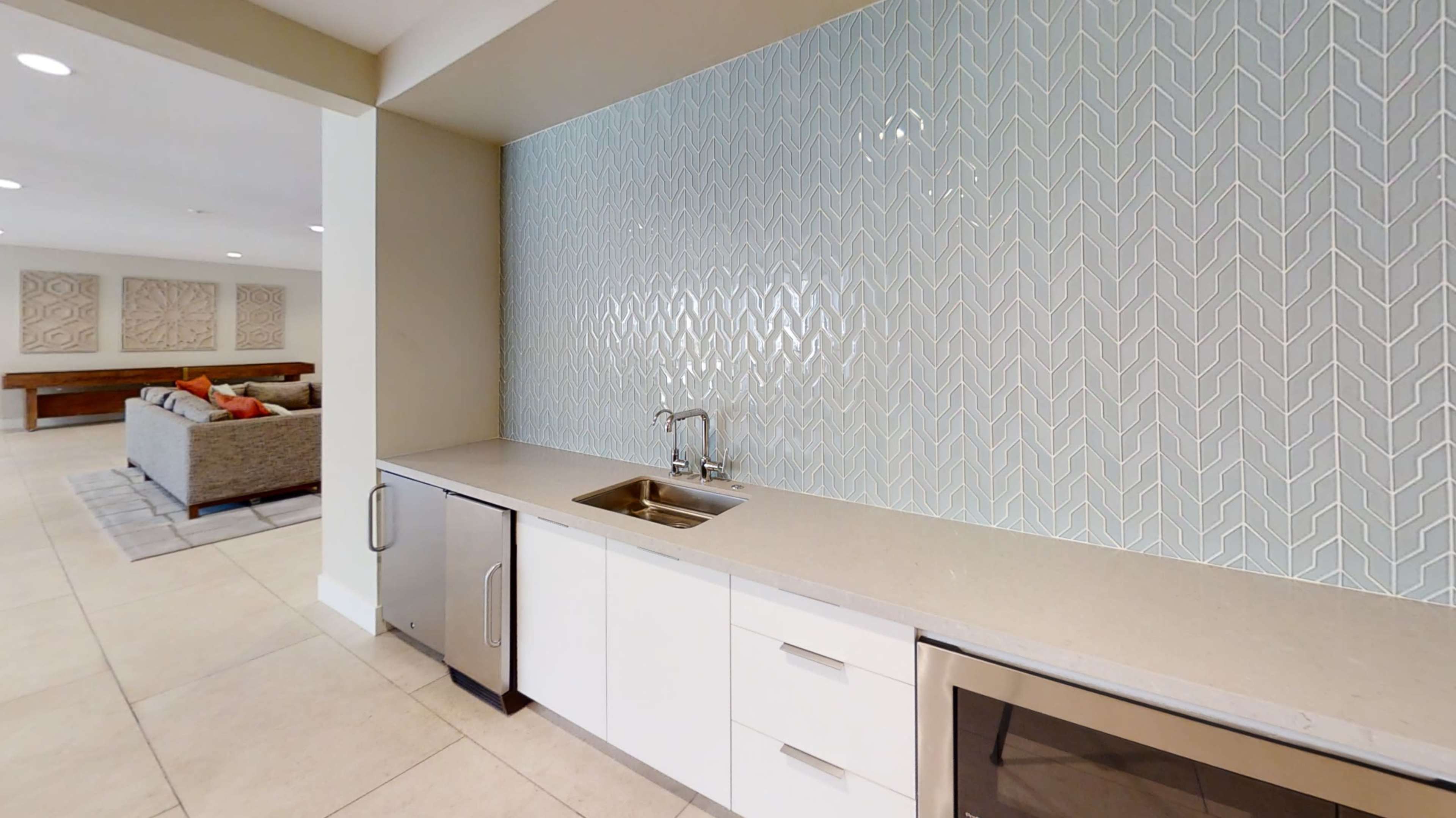 The image shows a modern kitchenette with a light blue herringbone-patterned backsplash, a stainless steel sink, and a microwave set into the cabinetry.