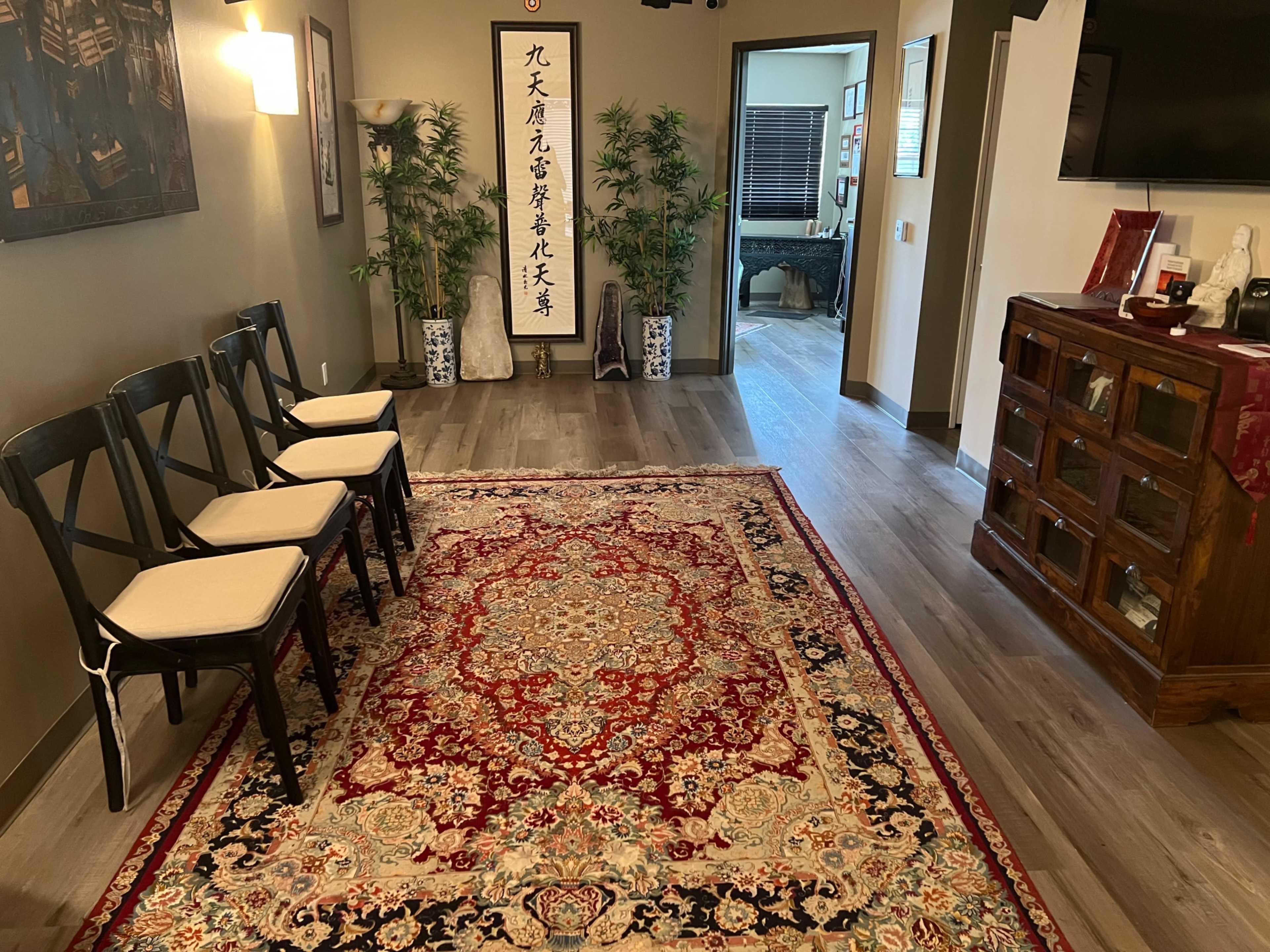 The image shows a neatly arranged waiting area with four chairs on a patterned rug, surrounded by potted plants and decorated walls, leading to a doorway marked by a Chinese calligraphy scroll.