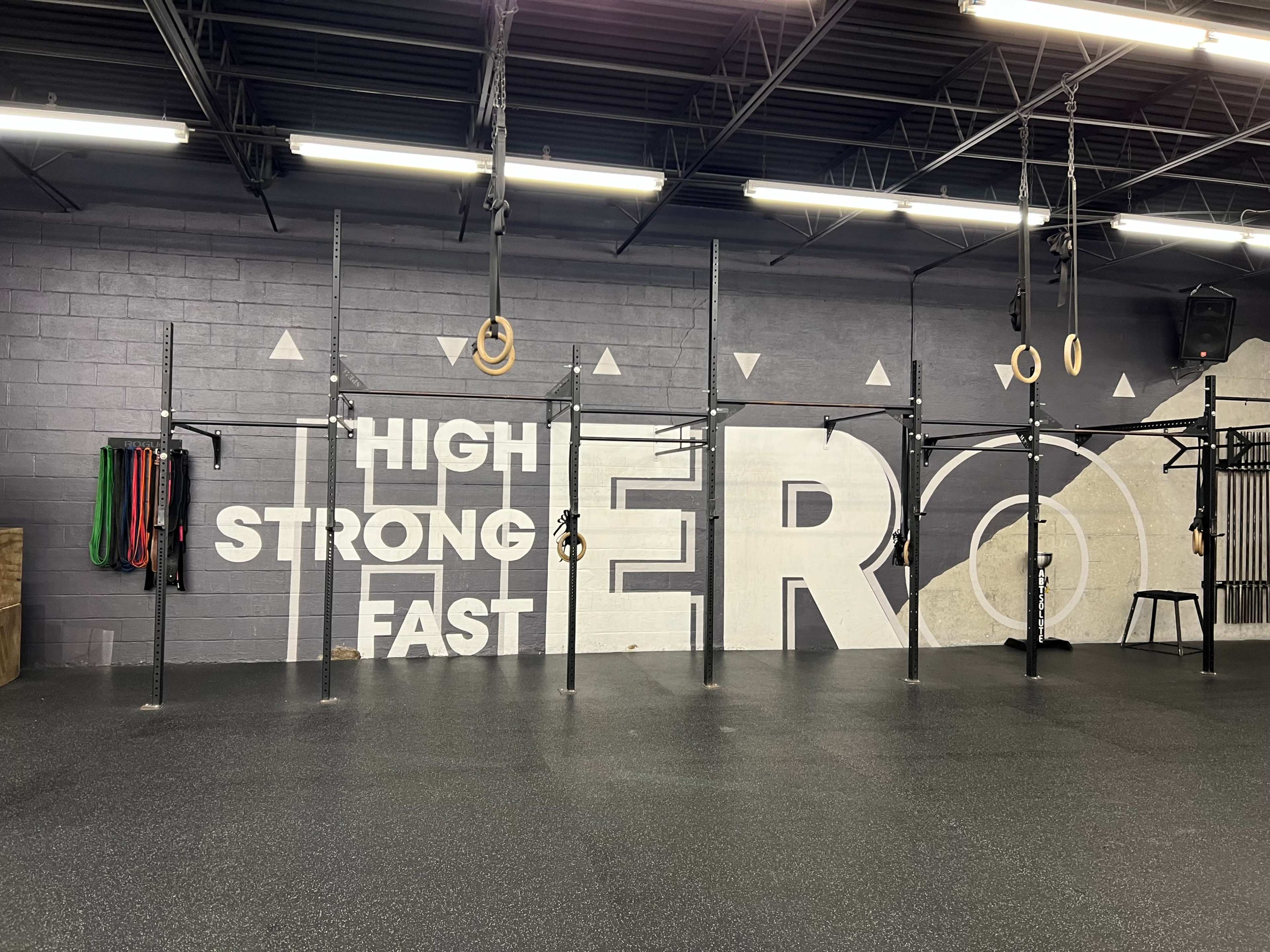 A gym interior with a black rubber floor, exercise equipment including rings and resistance bands, and a large mural featuring the words "HIGH STRONG FAST HERO."