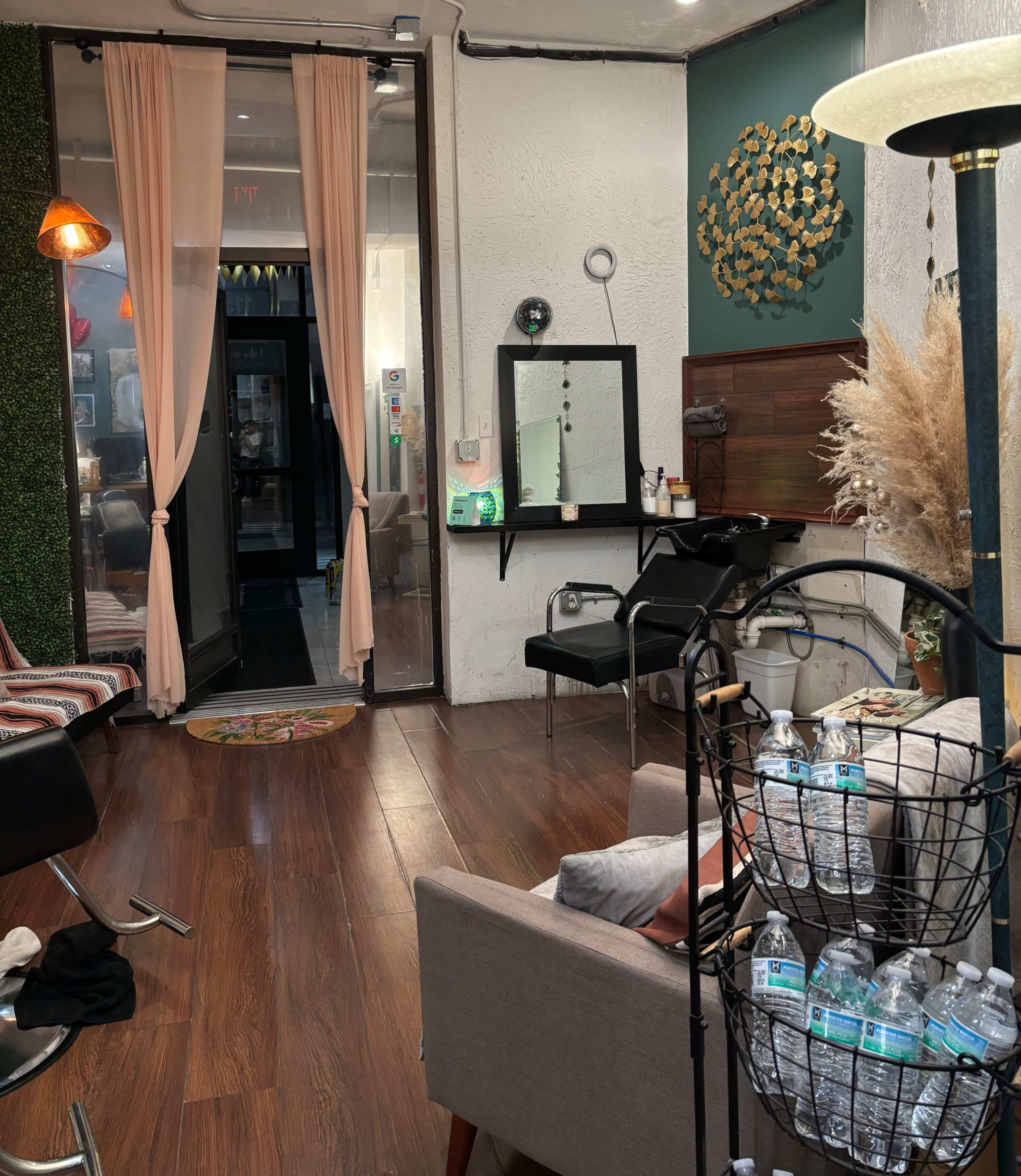 A salon interior with wooden flooring, chairs, a mirror, and a decorative wall, along with curtains and a basket of water bottles.