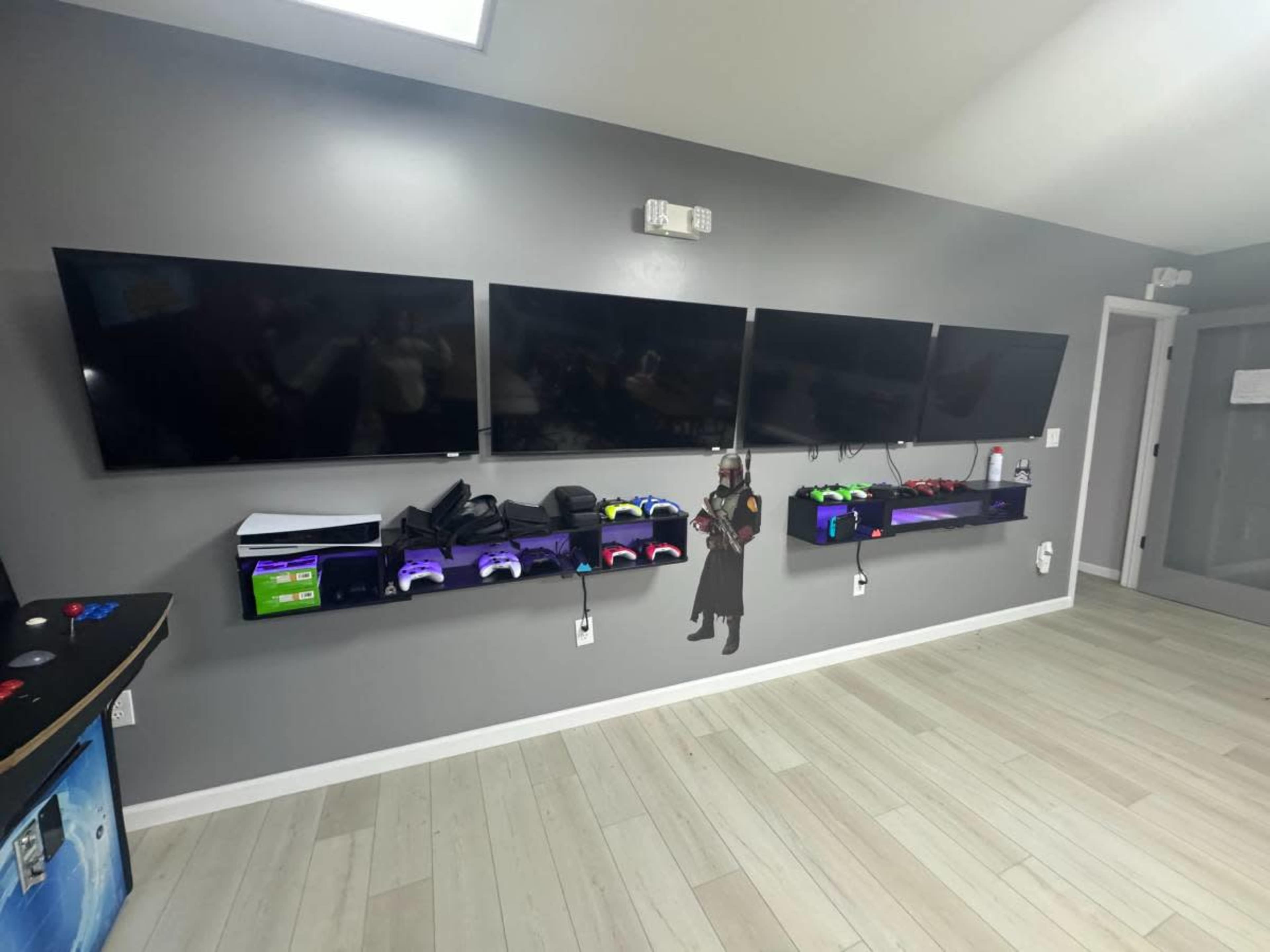 The image shows a wall with four mounted flat-screen televisions and a shelf below displaying various gaming controllers.