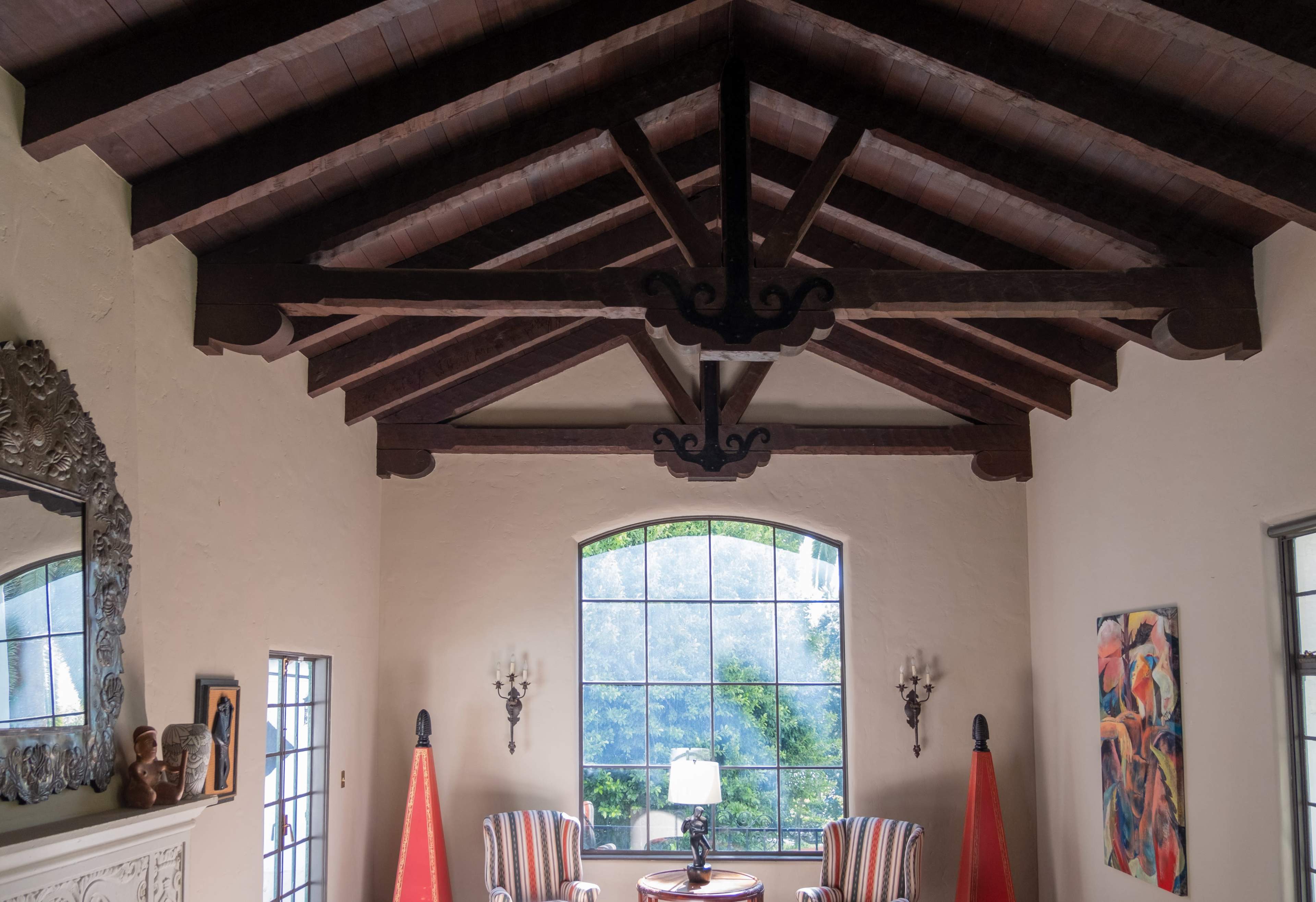 The interior of a room features a high vaulted ceiling with exposed wooden beams, large windows, and two striped armchairs beside a round table.