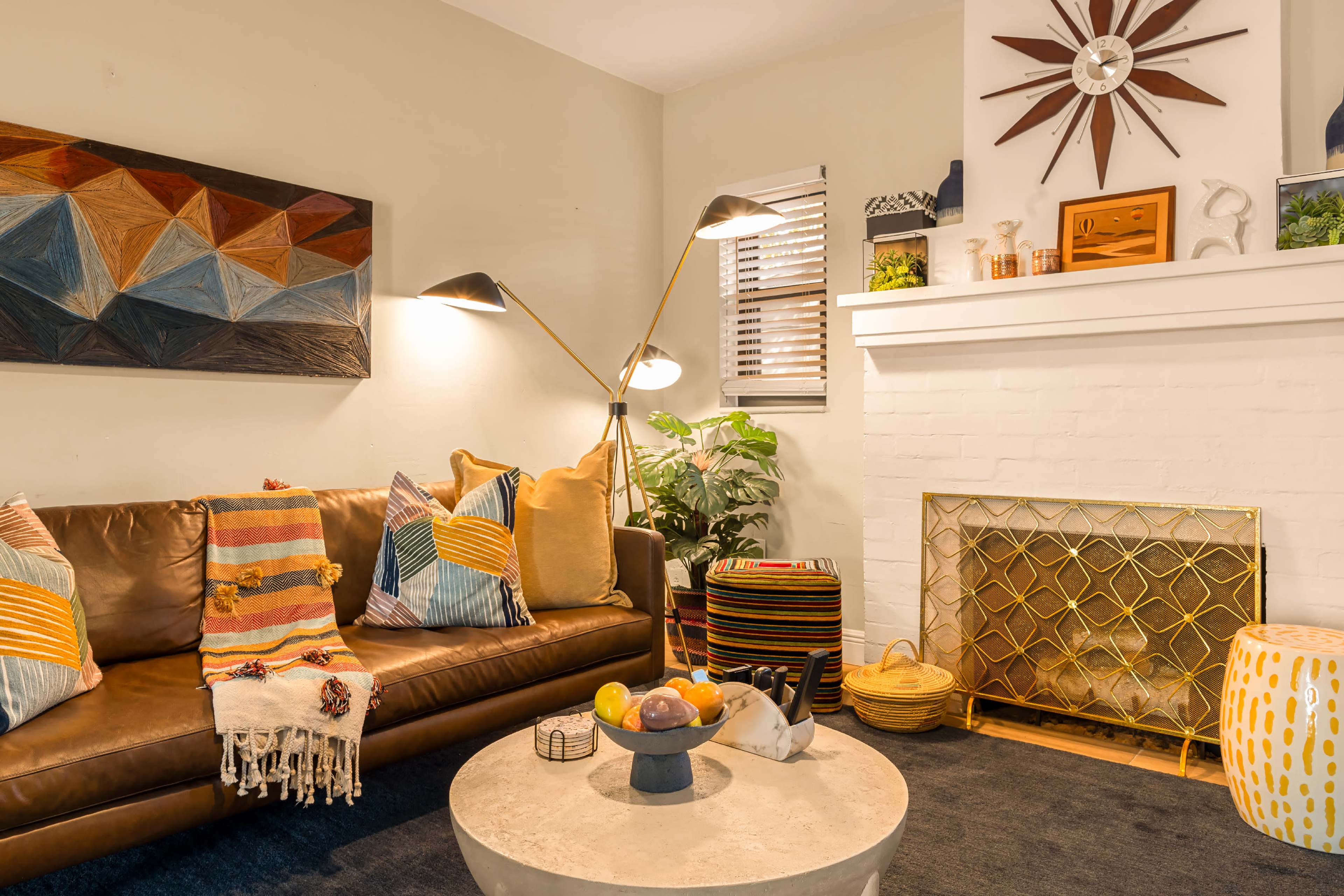 The image shows a cozy living room with a brown sofa adorned with colorful throw pillows, a round coffee table, and a white fireplace with decorative items.