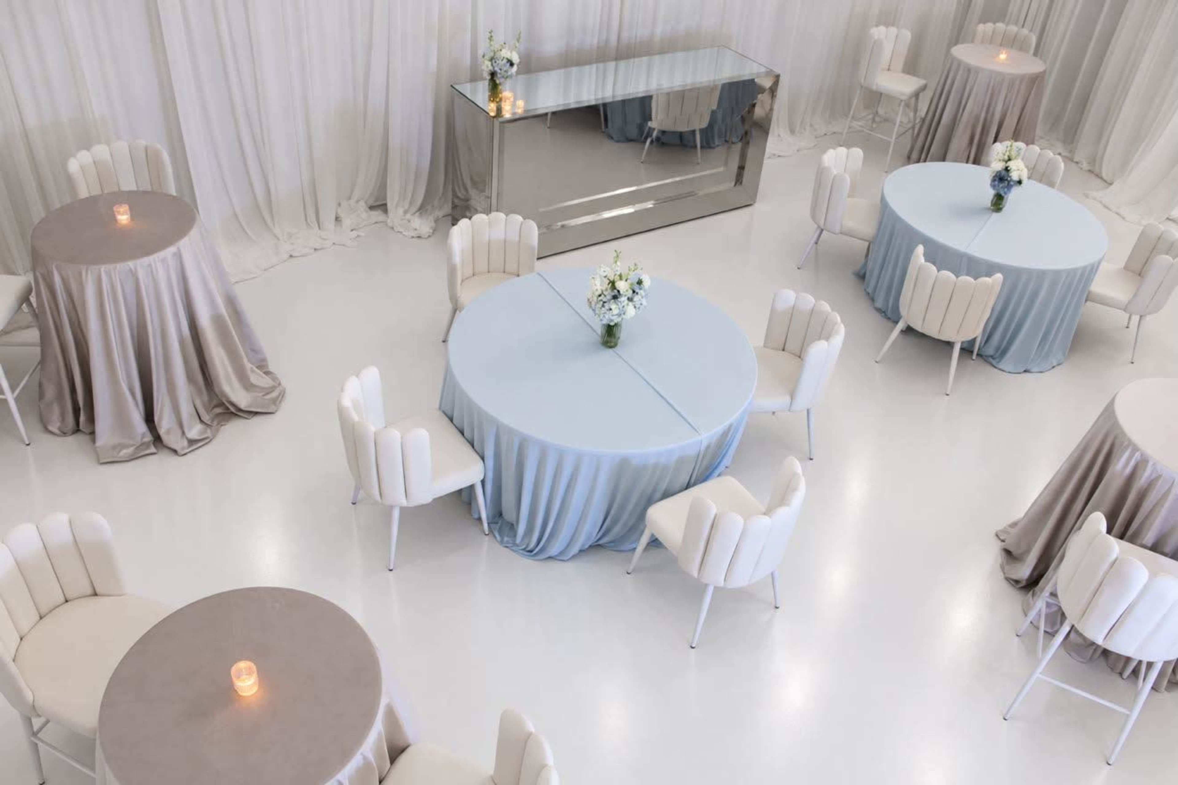 The image shows an elegantly arranged indoor space with round tables covered in light blue and beige tablecloths, accompanied by white chairs and a mirrored display table, illuminated by soft lighting.