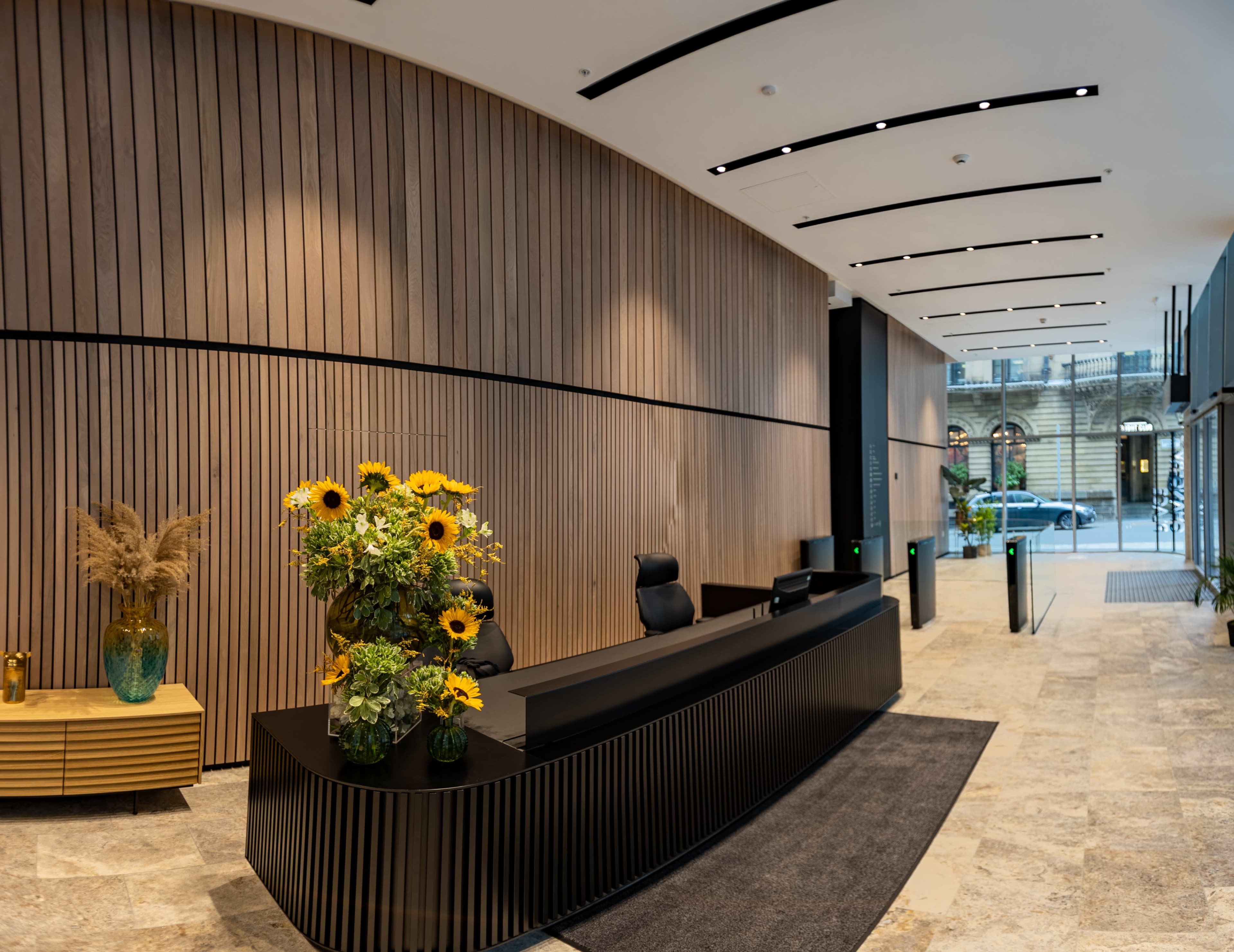 A modern reception area with a wooden wall, a sleek black reception desk, and a colorful arrangement of sunflowers on top.