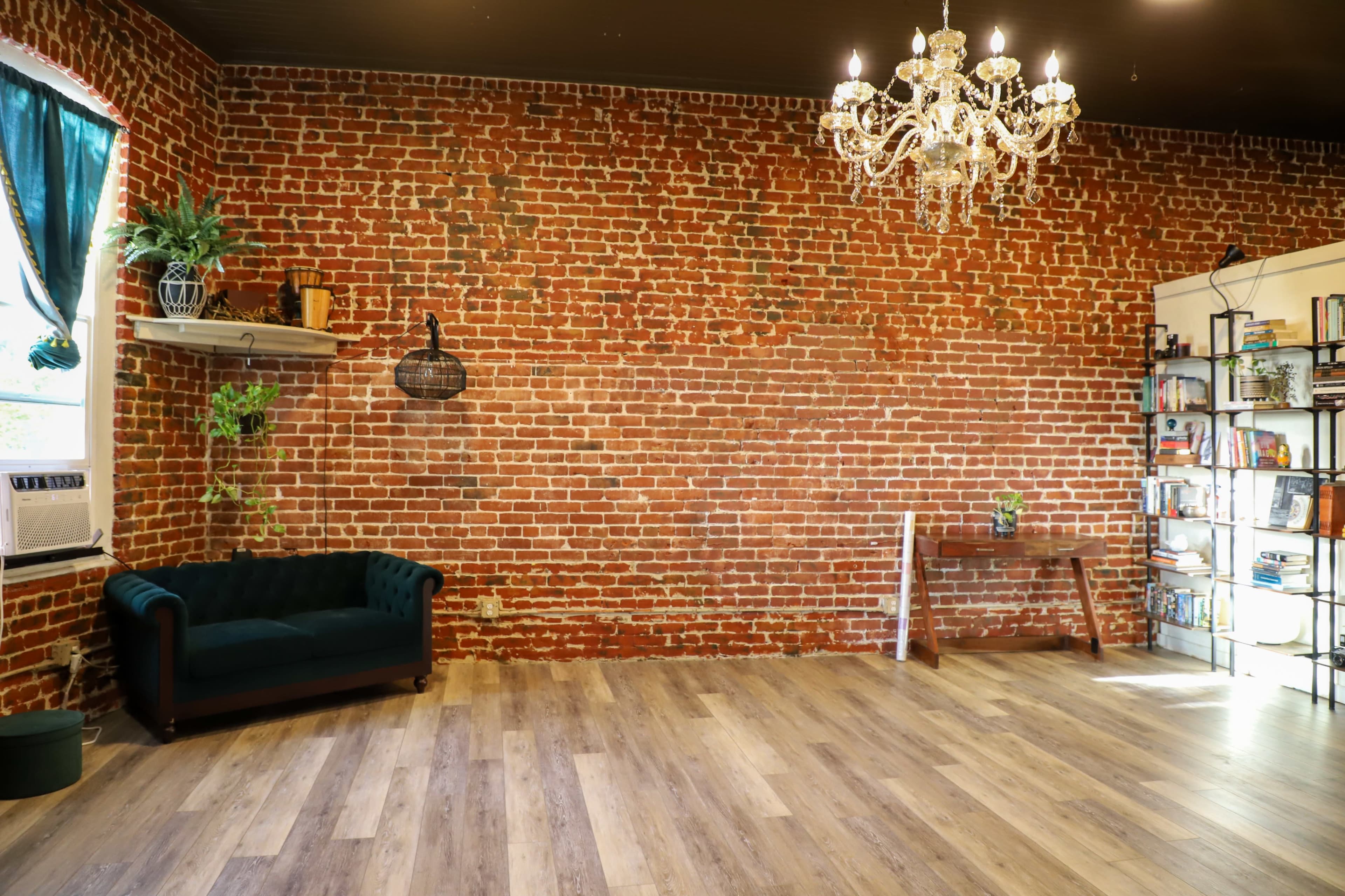 The room features a brick wall, a teal couch, a wooden table, a bookshelf filled with books, and a chandelier overhead.