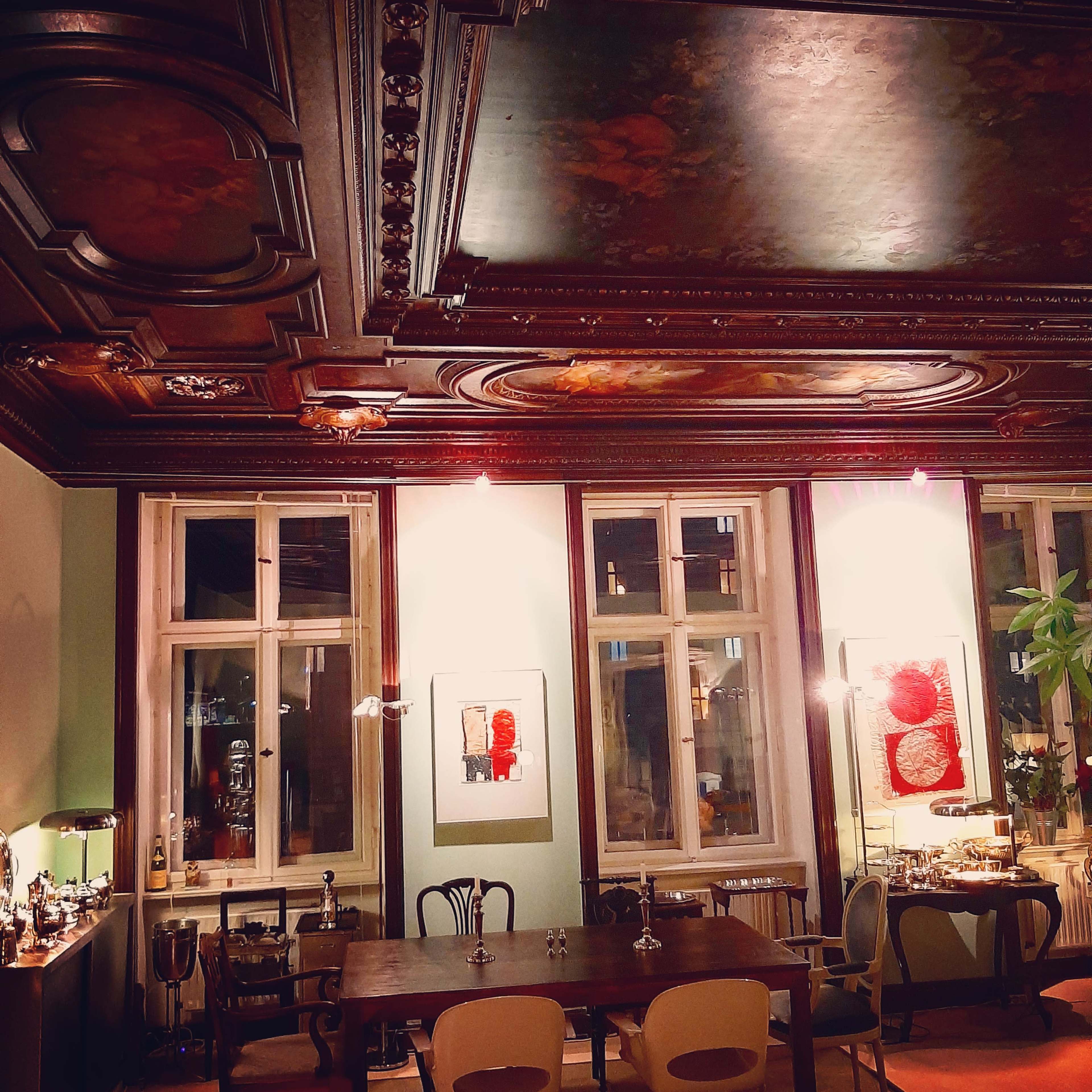 A warmly lit dining area with a wooden table, elegant ceiling details, and large windows revealing wall art and various table settings.