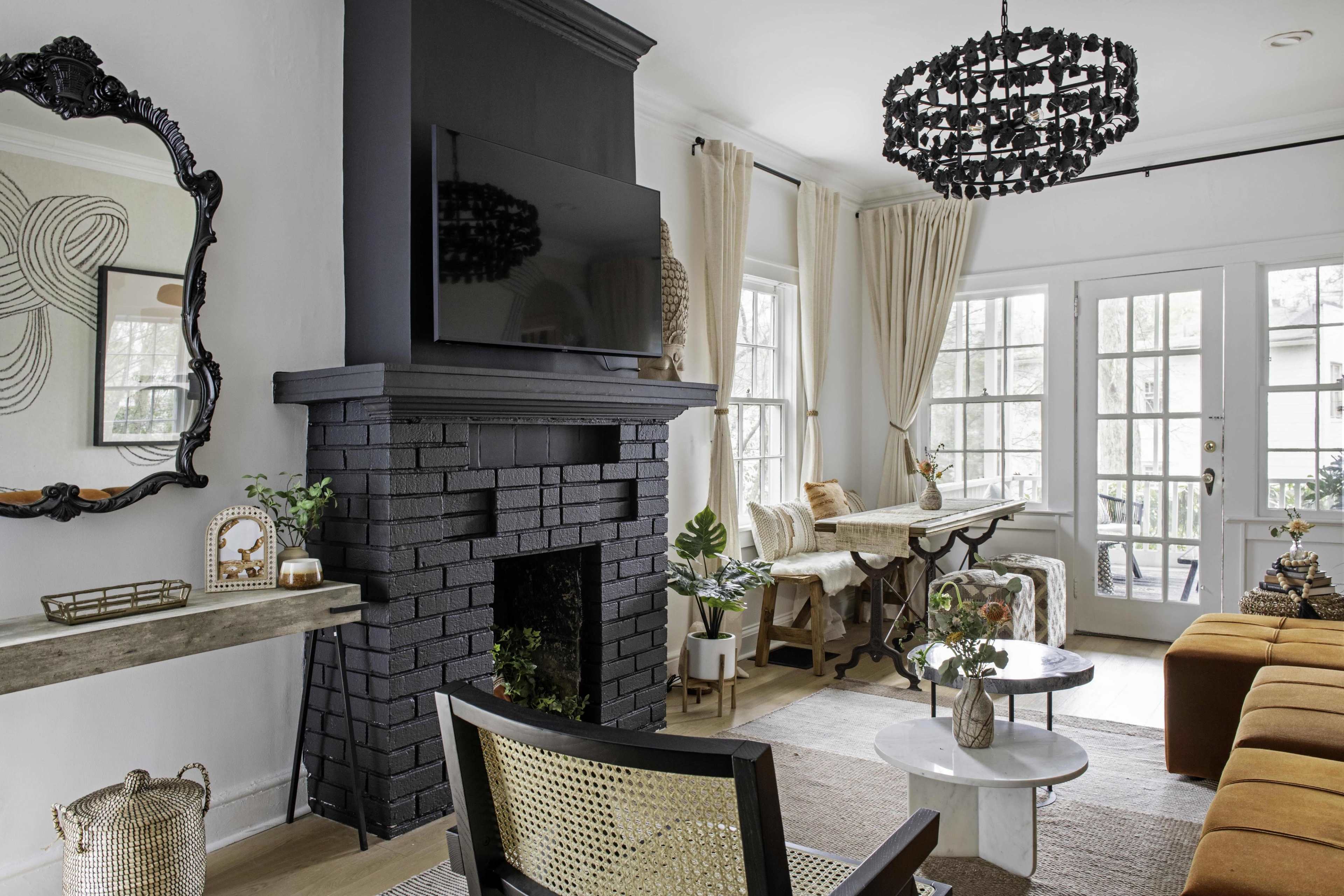 A modern living room features a black fireplace with a mounted television, a round coffee table, and a dining area with large windows, dressed with light curtains.