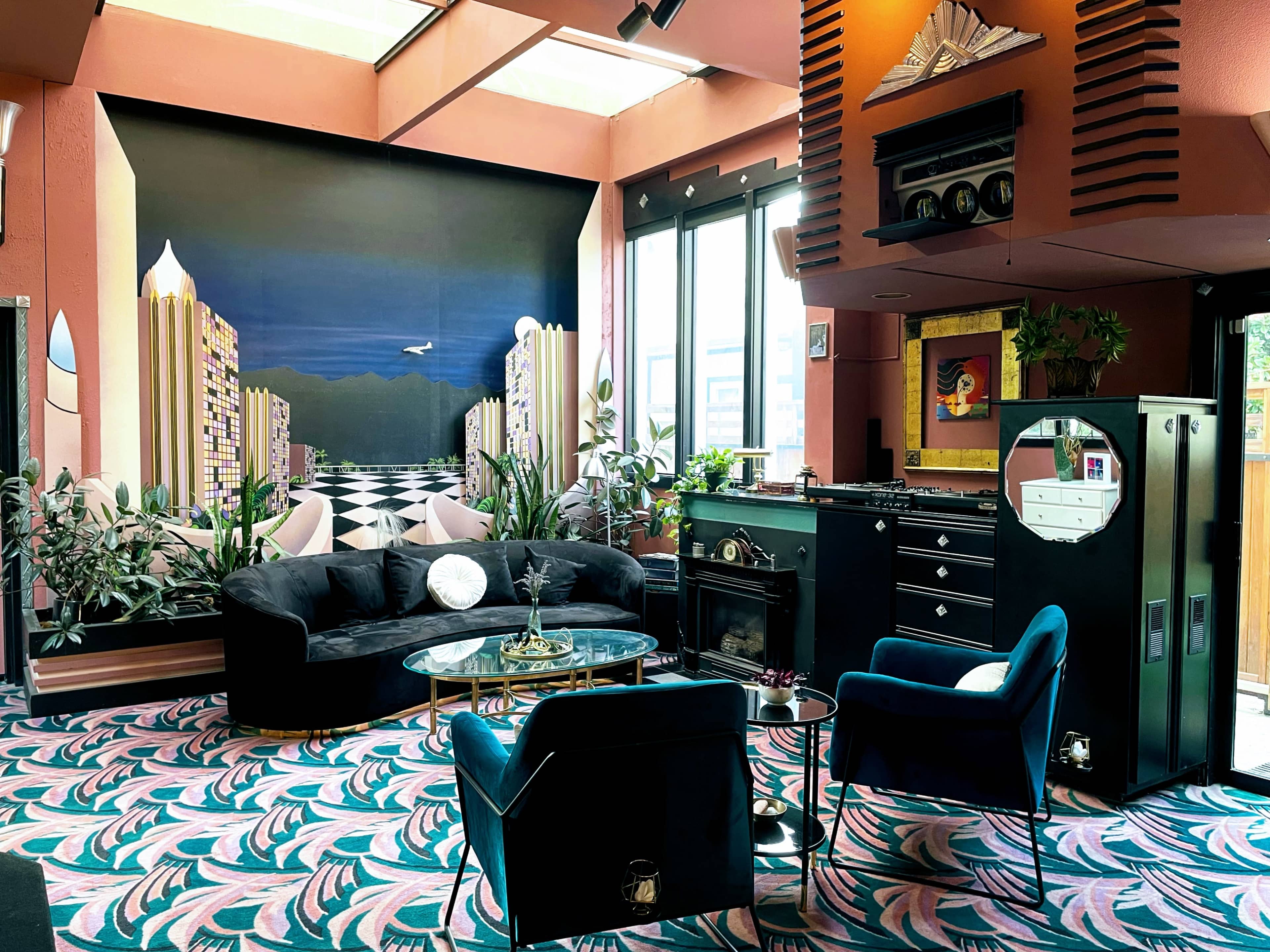 The scene features a stylish lounge area with a dark-colored couch and armchairs, surrounded by large windows and decorated with potted plants and a vibrant patterned carpet.