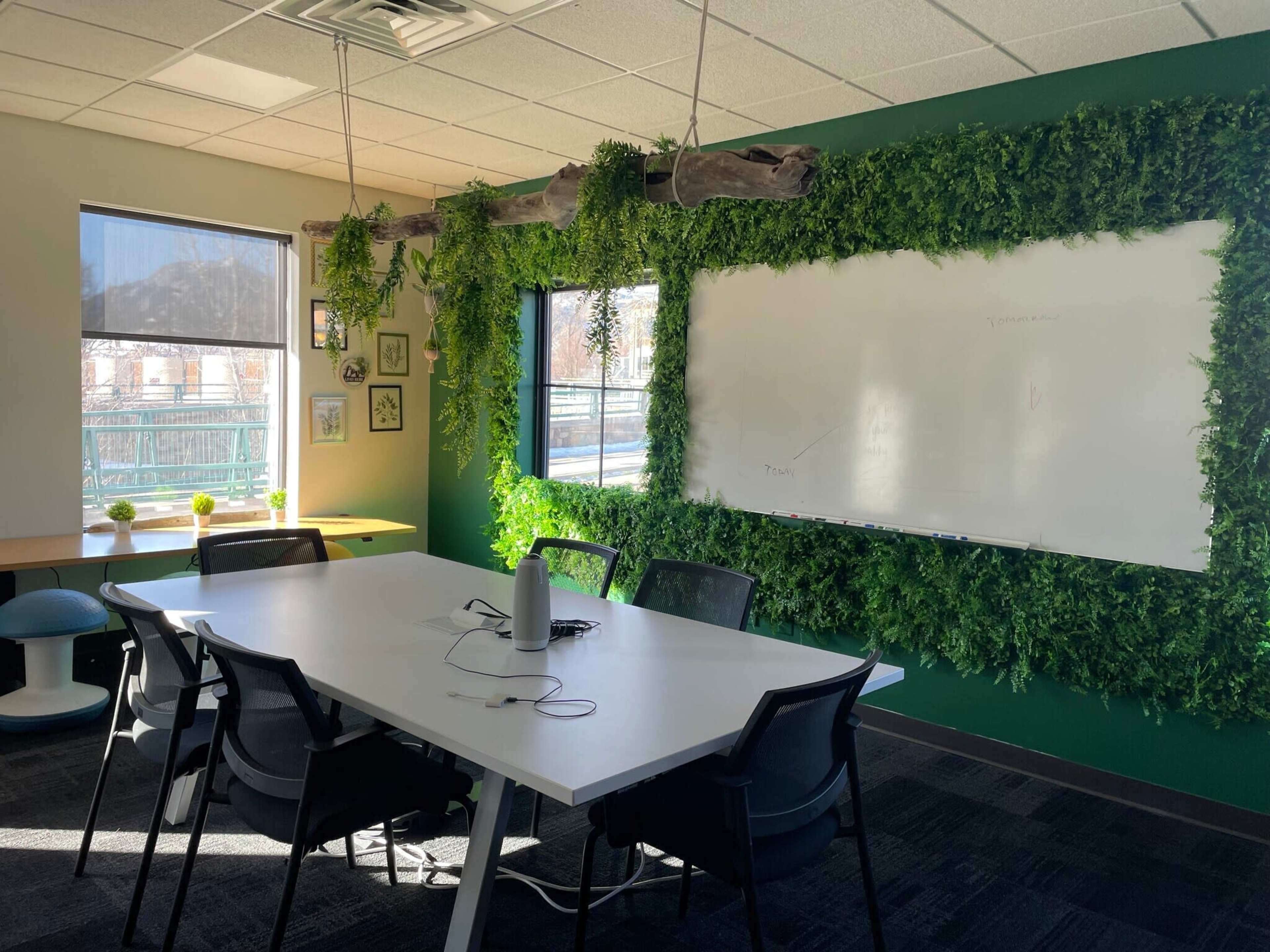 The image depicts a conference room adorned with green wall plants, featuring a whiteboard and a table surrounded by chairs.