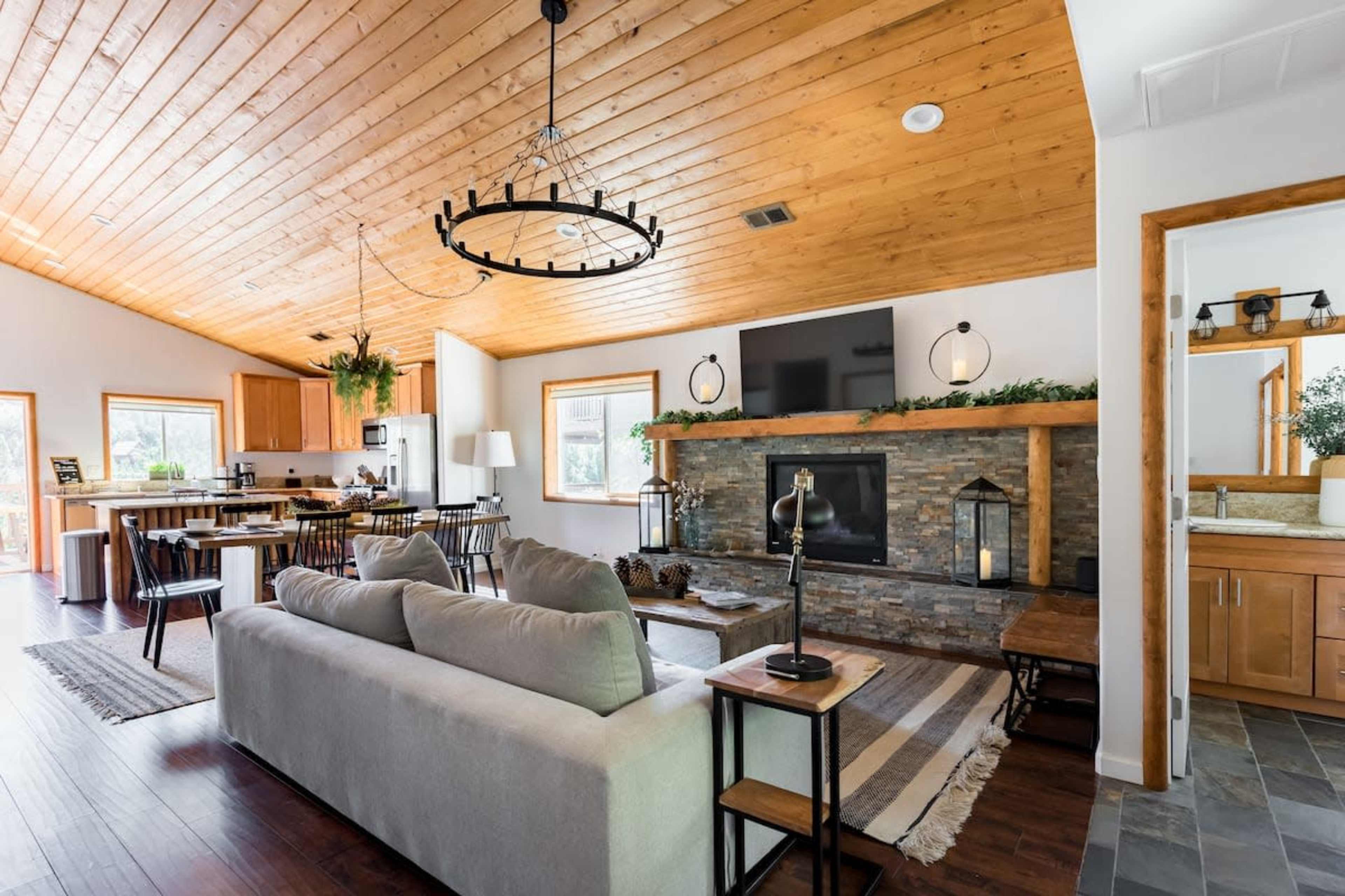 A spacious living area features a wooden ceiling, a stone fireplace, and modern furnishings, connecting to an open kitchen.