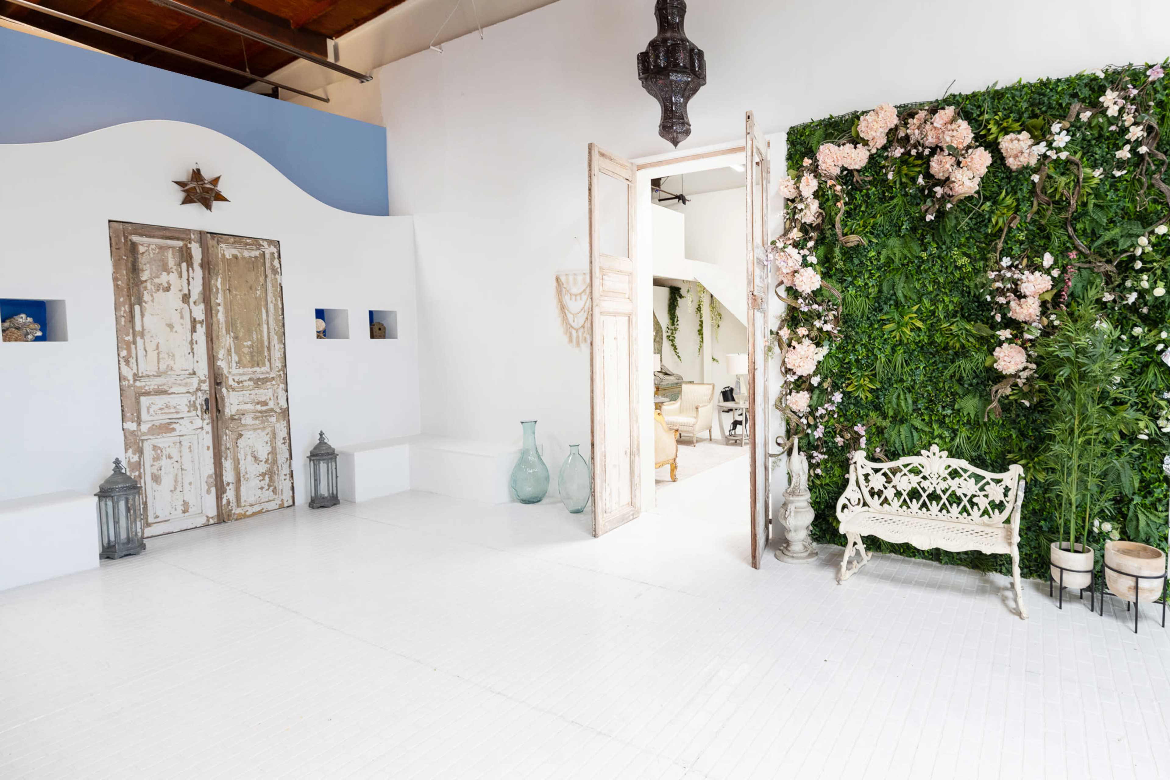 The image shows a bright, airy interior featuring rustic wooden doors, a green wall adorned with flowers, and a decorative white bench.