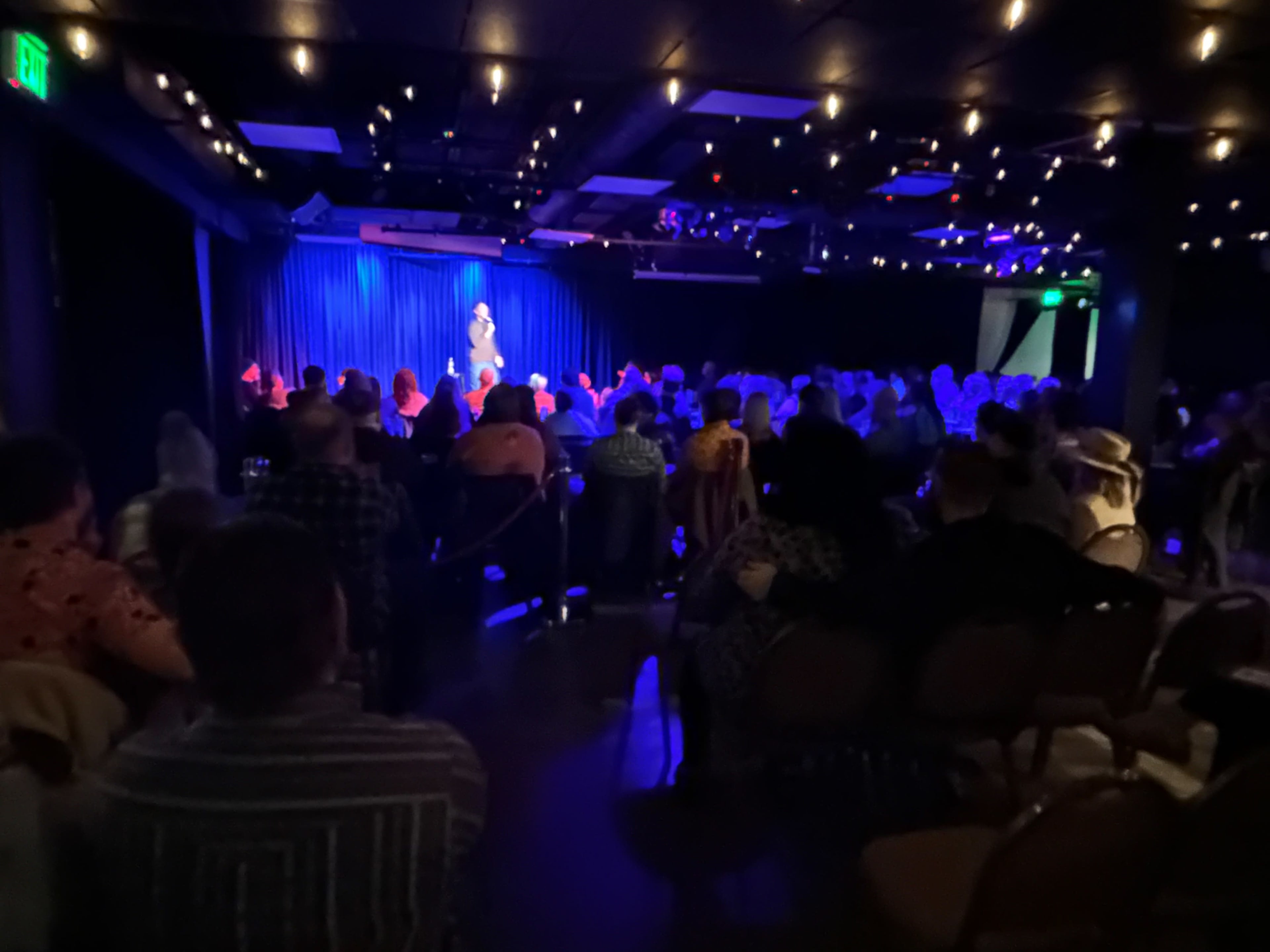 A comedian performs on stage at a dimly lit comedy club, with an audience seated in front.