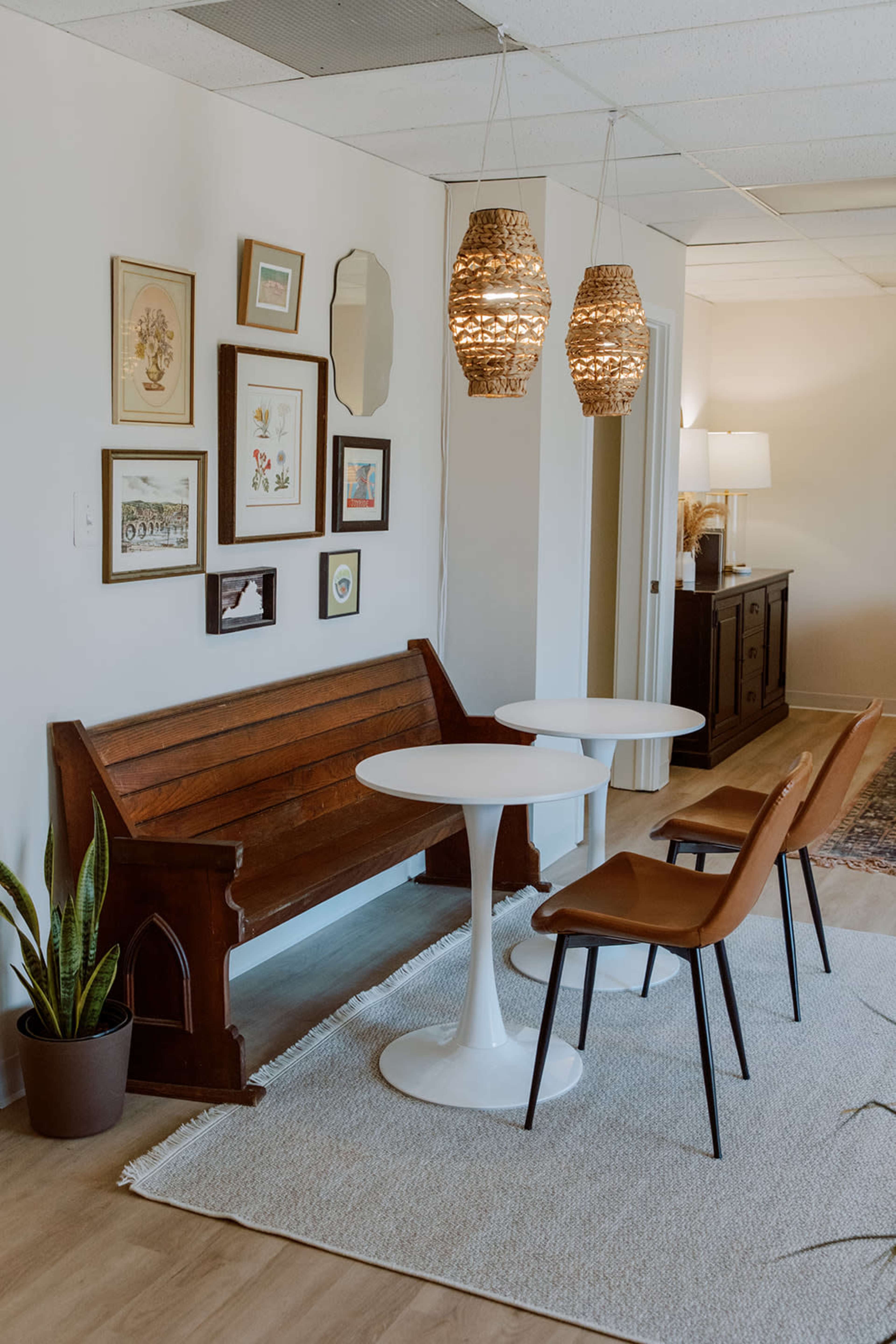 A wooden bench is positioned against a wall decorated with framed artwork, accompanied by two dining chairs and a round table, with woven pendant lights hanging above.