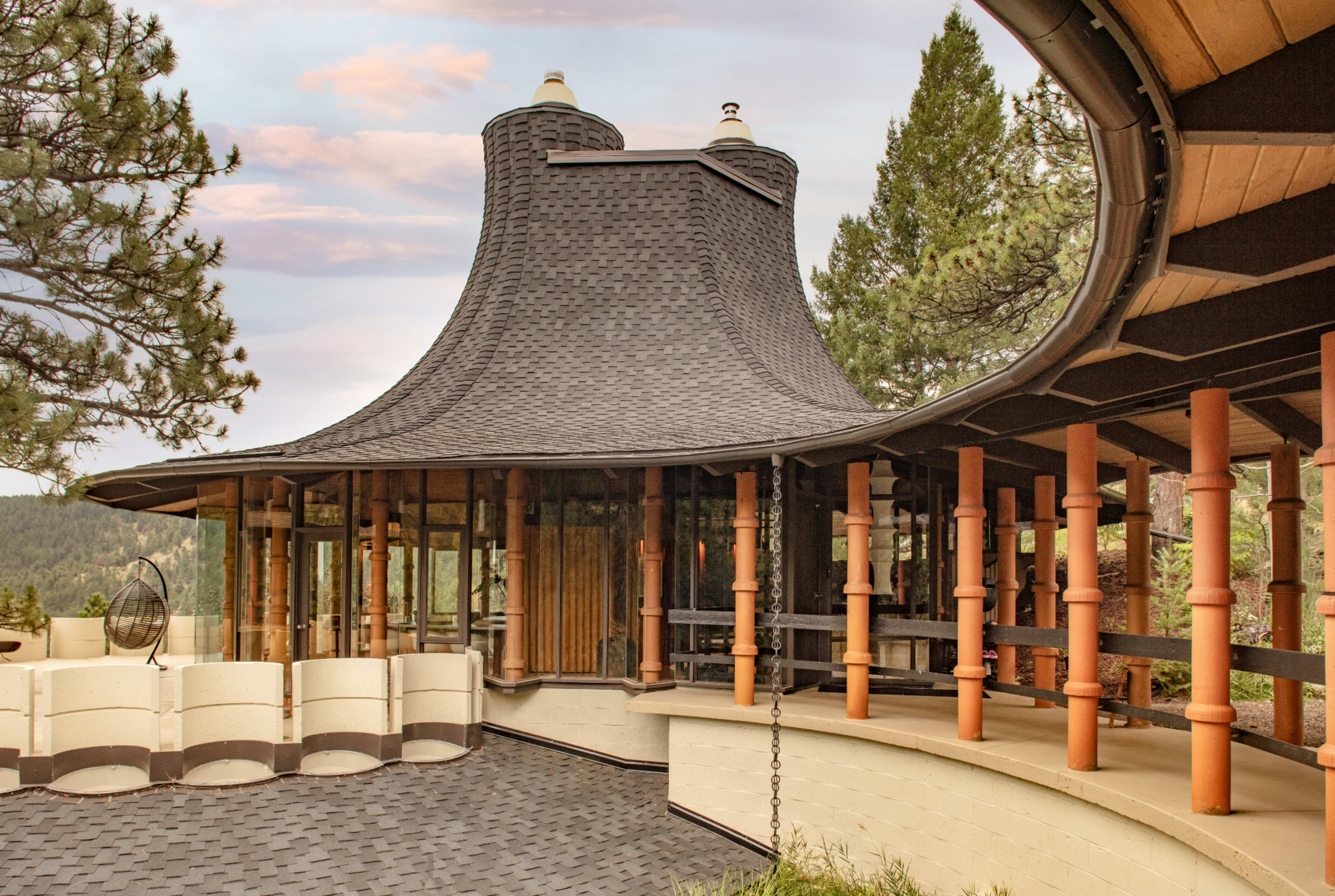 The image showcases a unique architectural structure with a curved roof and wooden support pillars, set amidst a natural landscape of trees and mountains.