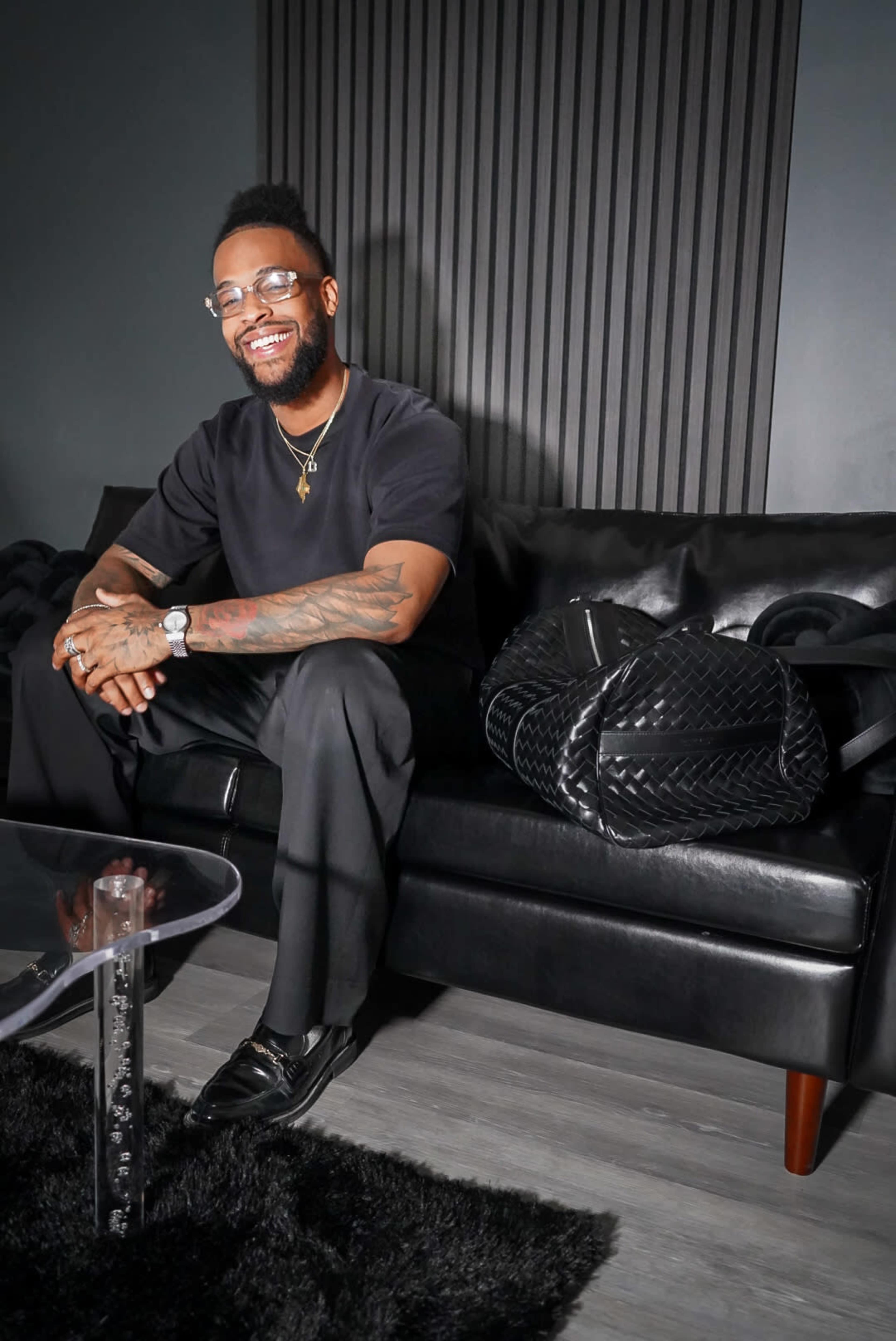 A man with glasses and tattoos smiles while sitting on a black leather couch next to a large black bag.