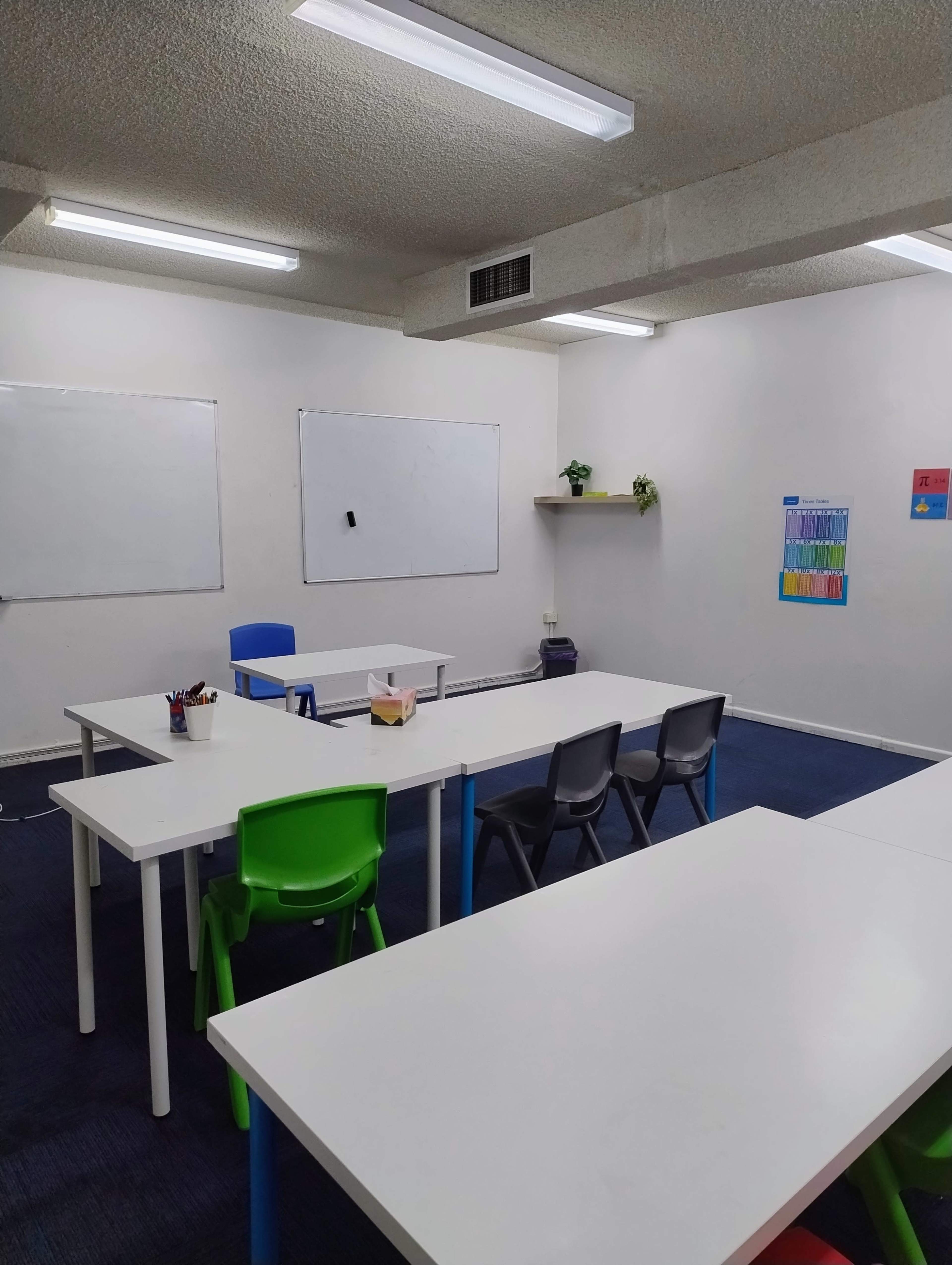 The image shows a classroom with several white tables and chairs arranged around the space, along with whiteboards and a plant on a shelf.
