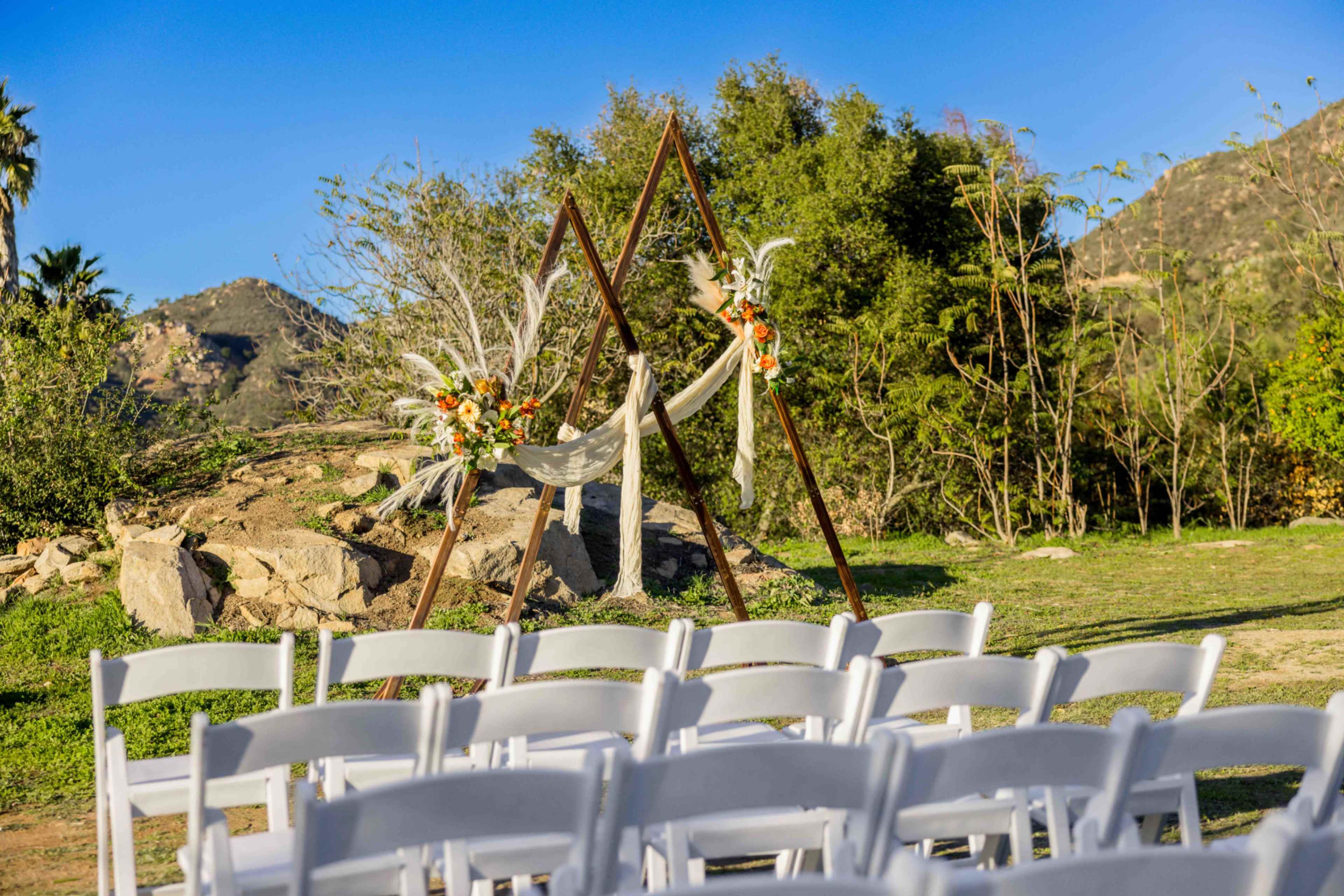The image shows an outdoor wedding setup featuring a floral arch with two triangular structures and white folding chairs arranged in front of it.