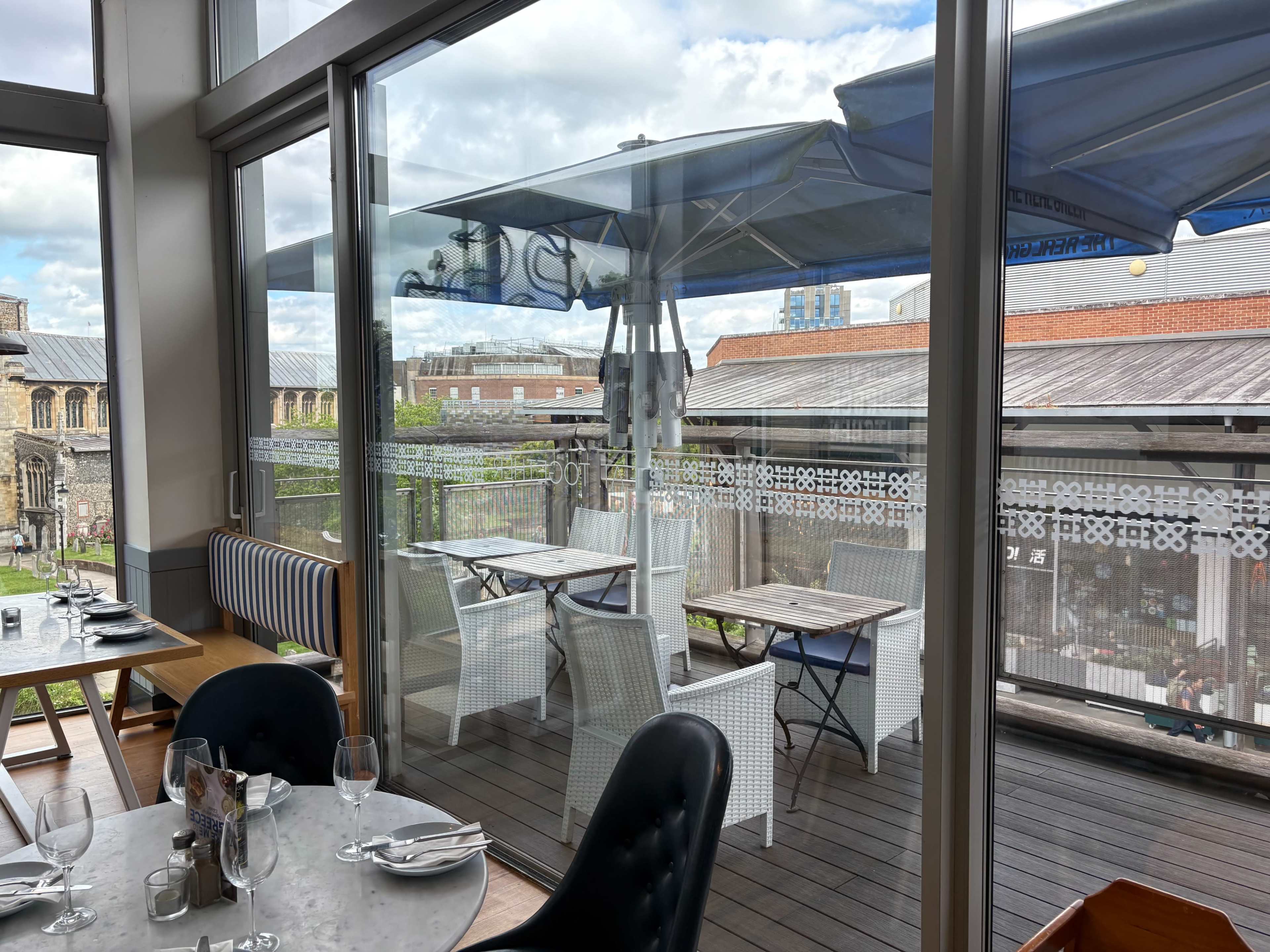 The image shows a restaurant interior with tables set for dining, overlooking a patio area featuring umbrellas and outdoor seating.