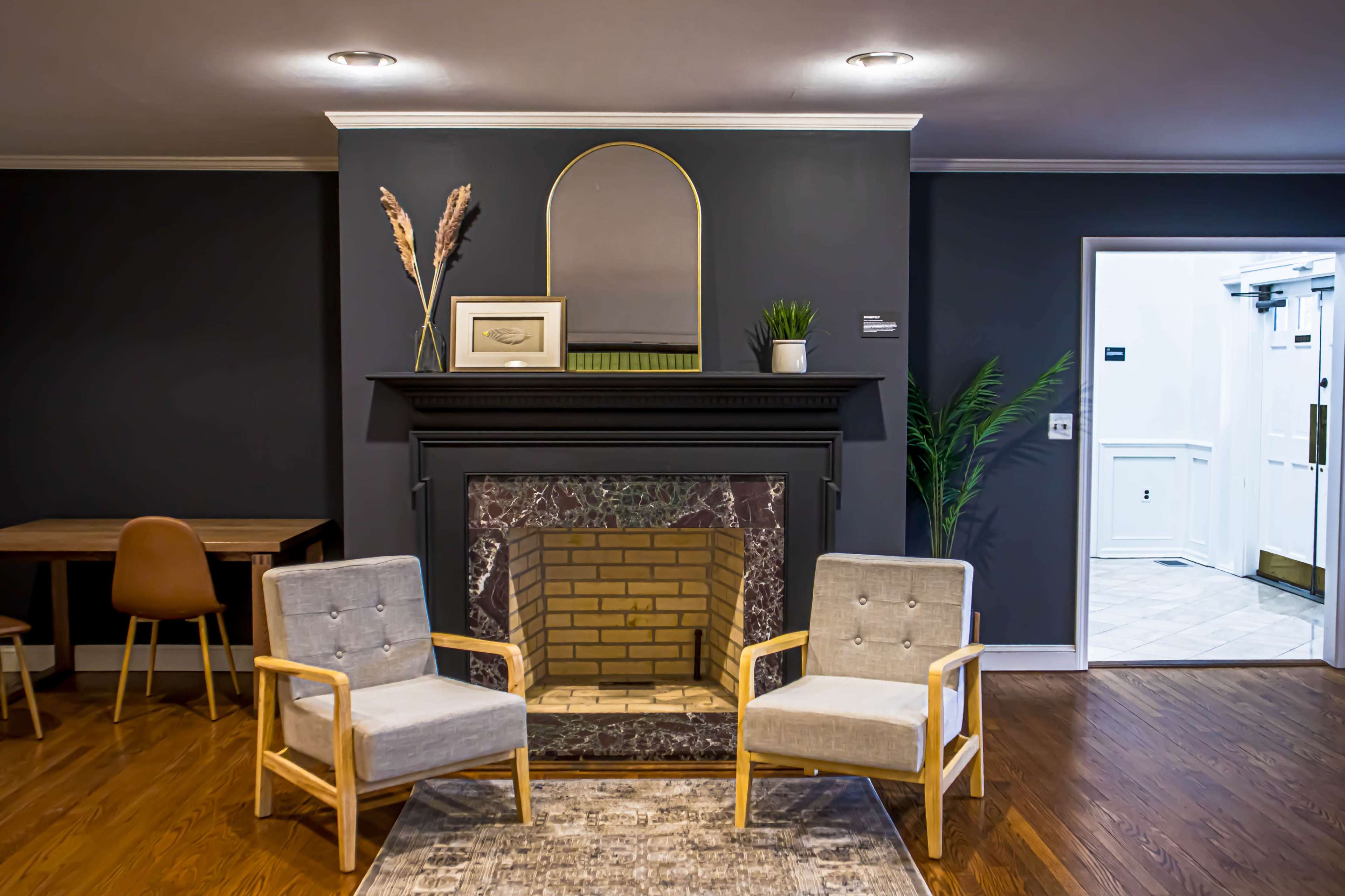 A modern living space featuring two gray chairs positioned in front of a brick fireplace, with a mirror and decorative plants above the mantel.