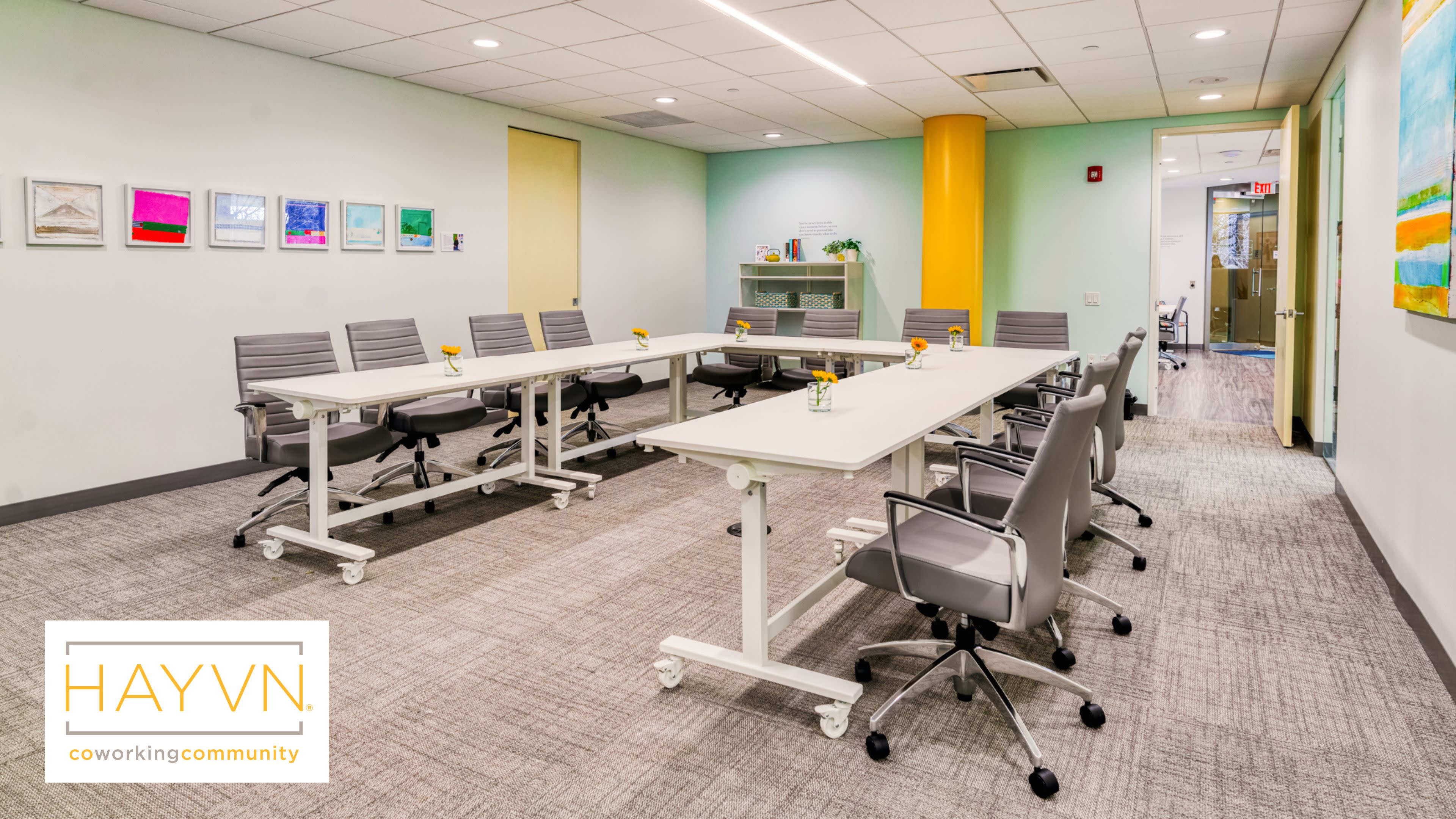 The image shows a well-lit conference room featuring a long white table surrounded by gray ergonomic chairs, with artwork on the yellow walls and a side shelf holding decorative items.