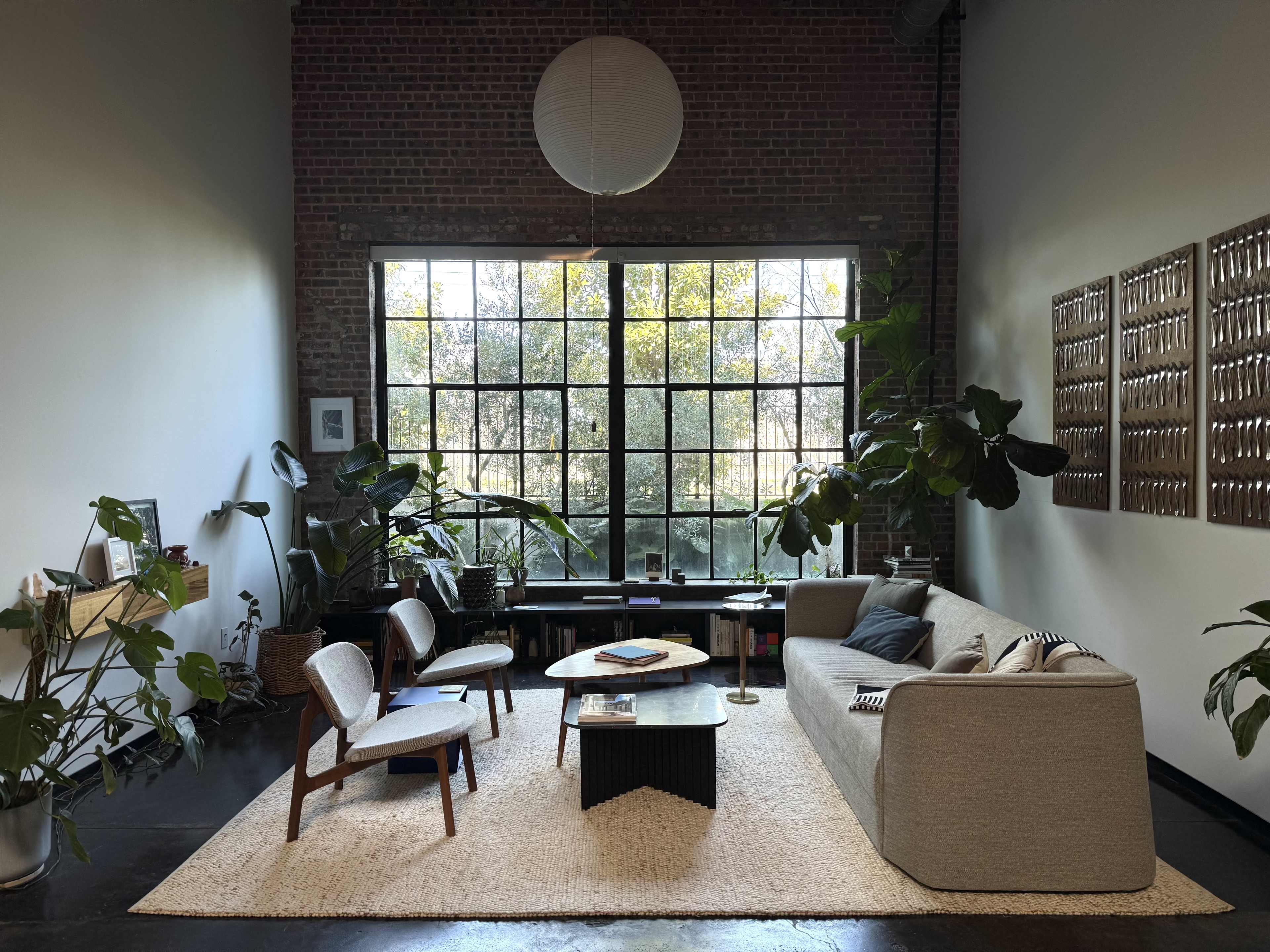 The image shows a cozy living room featuring a large window, a light-colored sofa, two wooden chairs, a coffee table, and several indoor plants.
