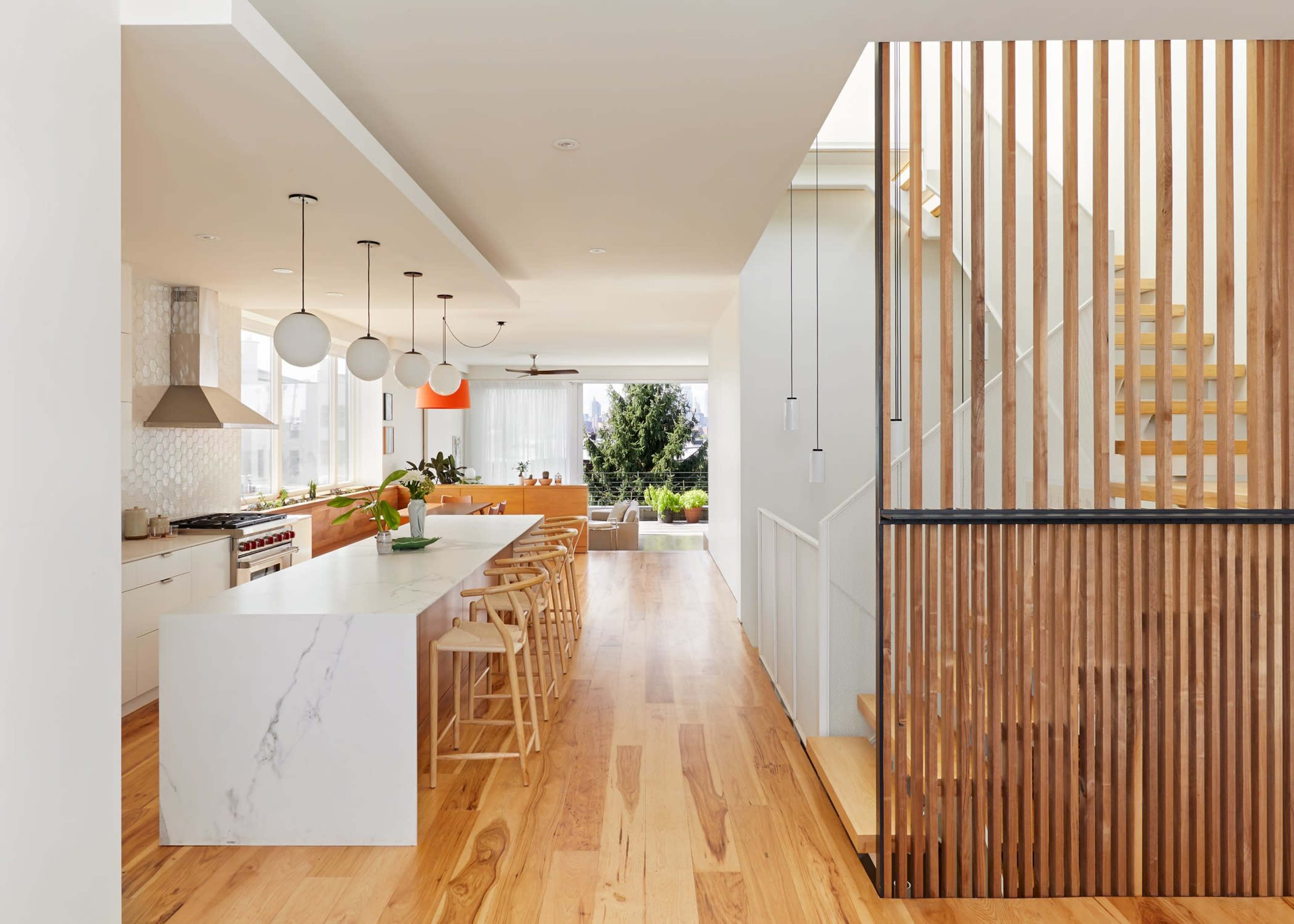 The image shows a modern kitchen and dining area with a long kitchen island, wooden flooring, and a staircase made of wood and metal.