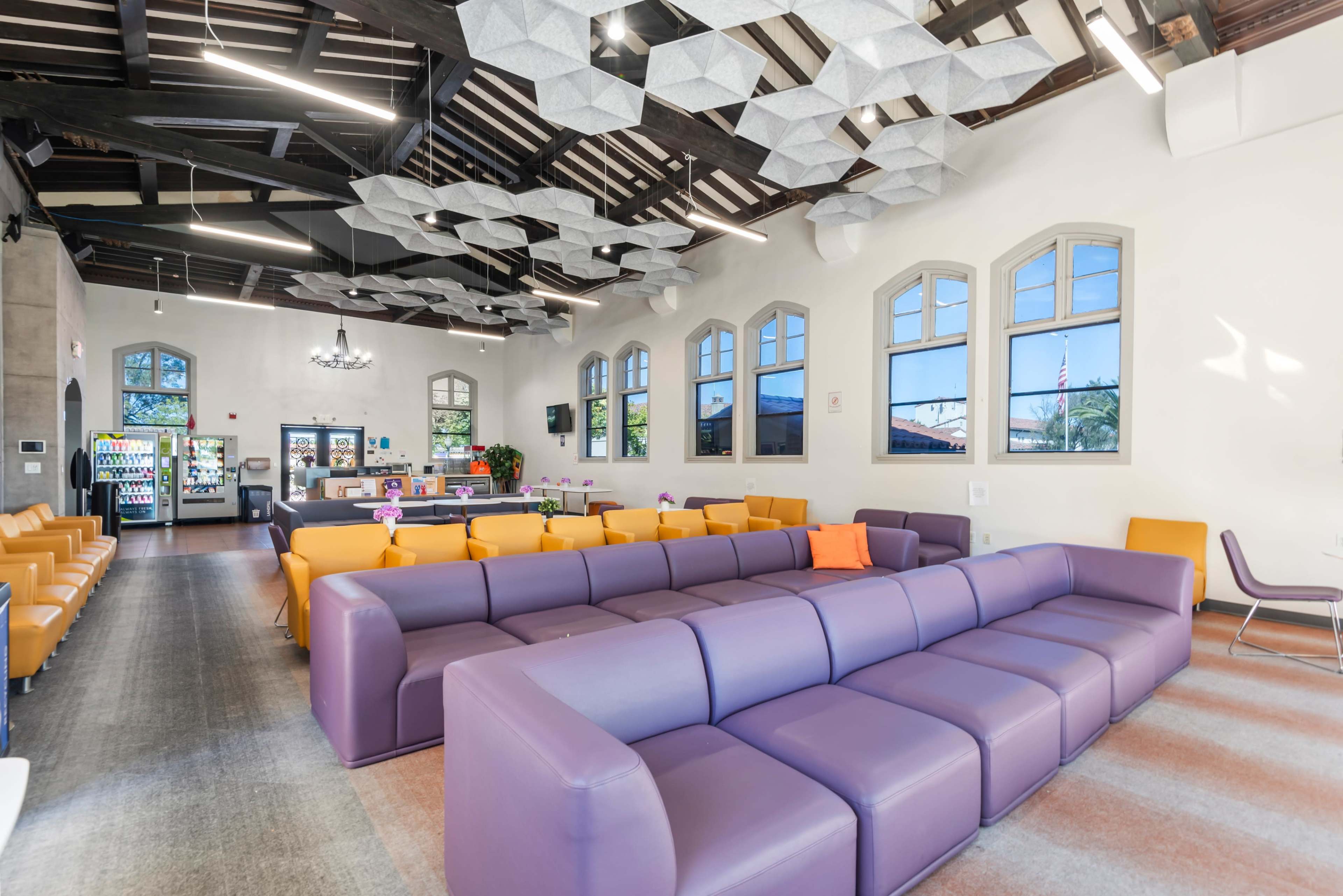 The image shows a spacious, modern lounge area with large windows, a mix of purple and orange seating, and contemporary decorative light fixtures on the ceiling.