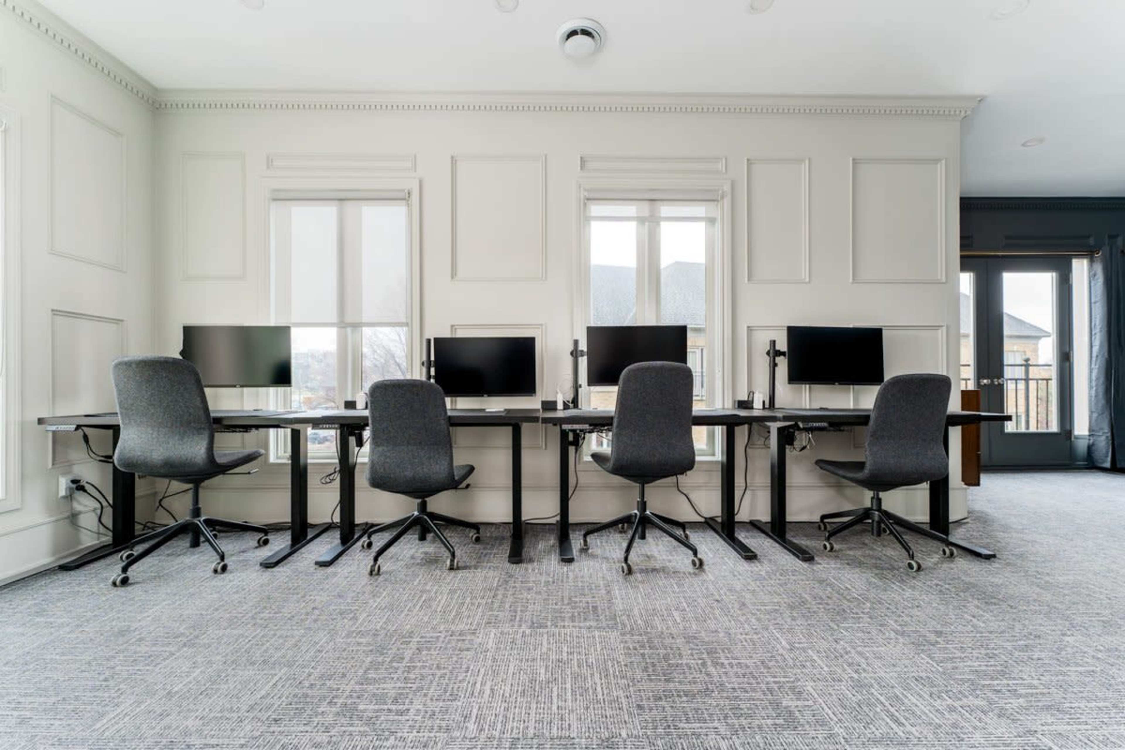 A modern office space with four ergonomic chairs and desks, each equipped with computer monitors, set against light-colored walls and large windows.