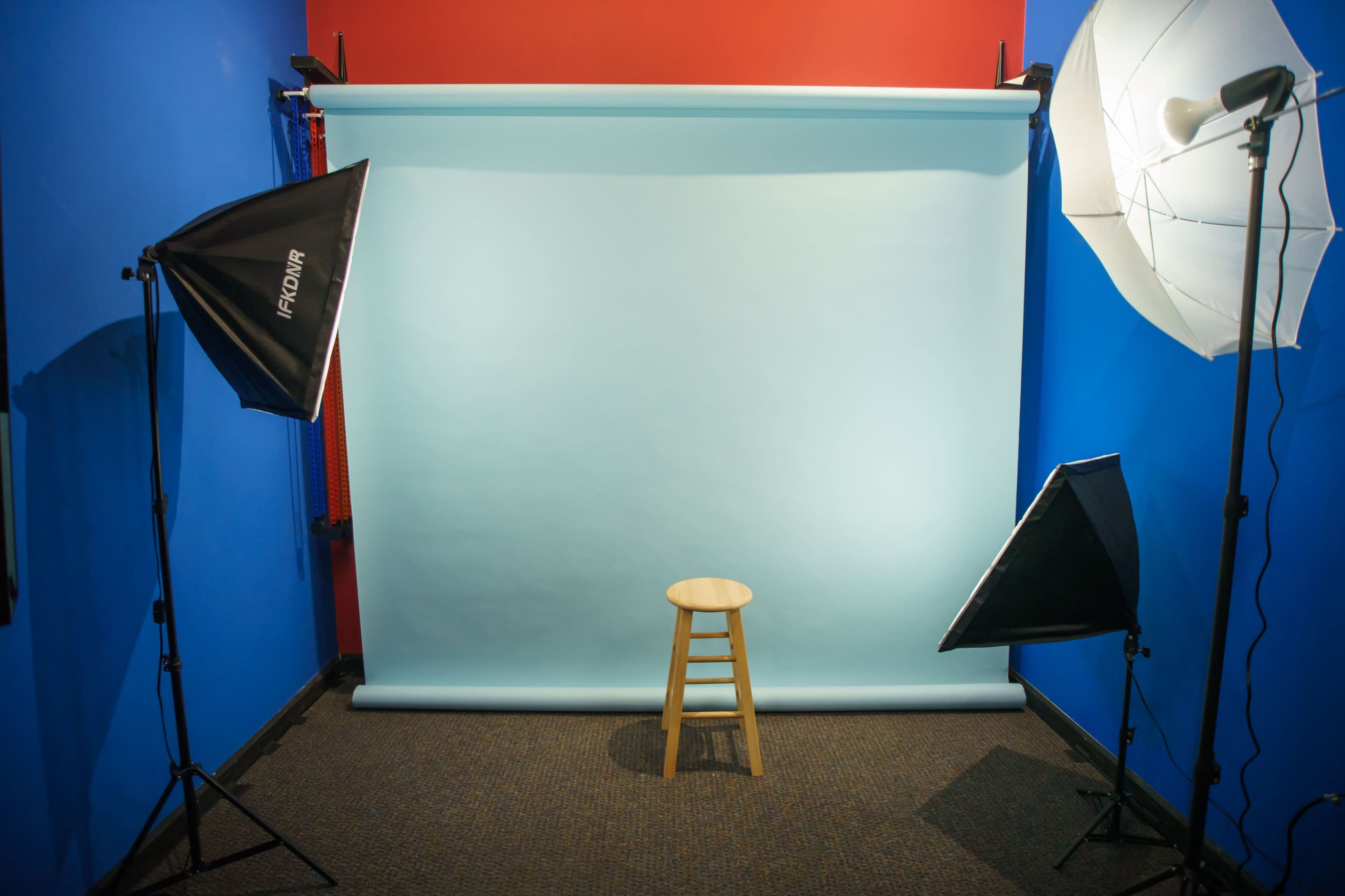 A photography studio with a light blue backdrop, a wooden stool in the center, and two softbox lights positioned on either side.