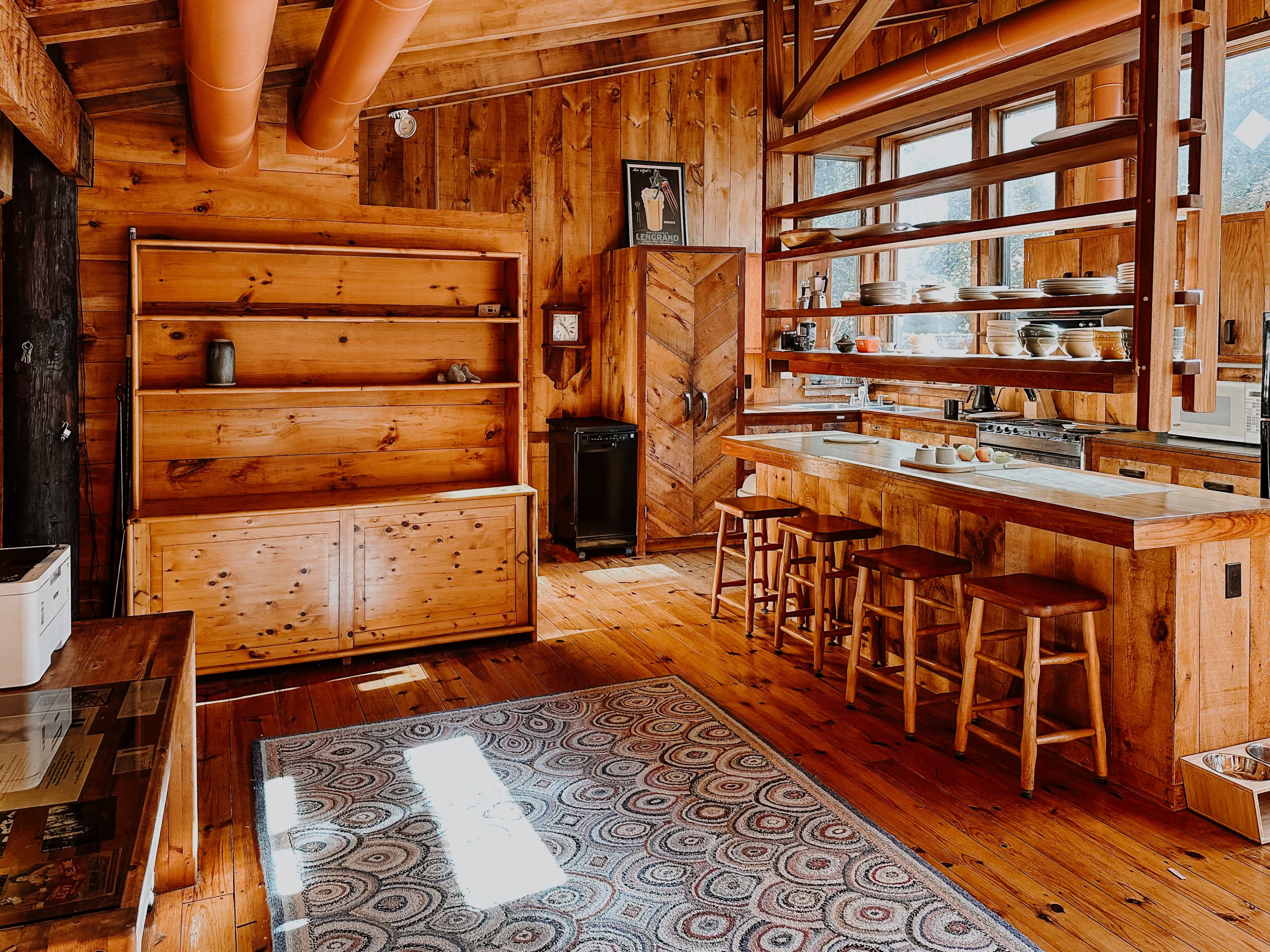 The image shows a rustic kitchen with wooden walls, shelving, a central island with barstools, and a large window allowing natural light.