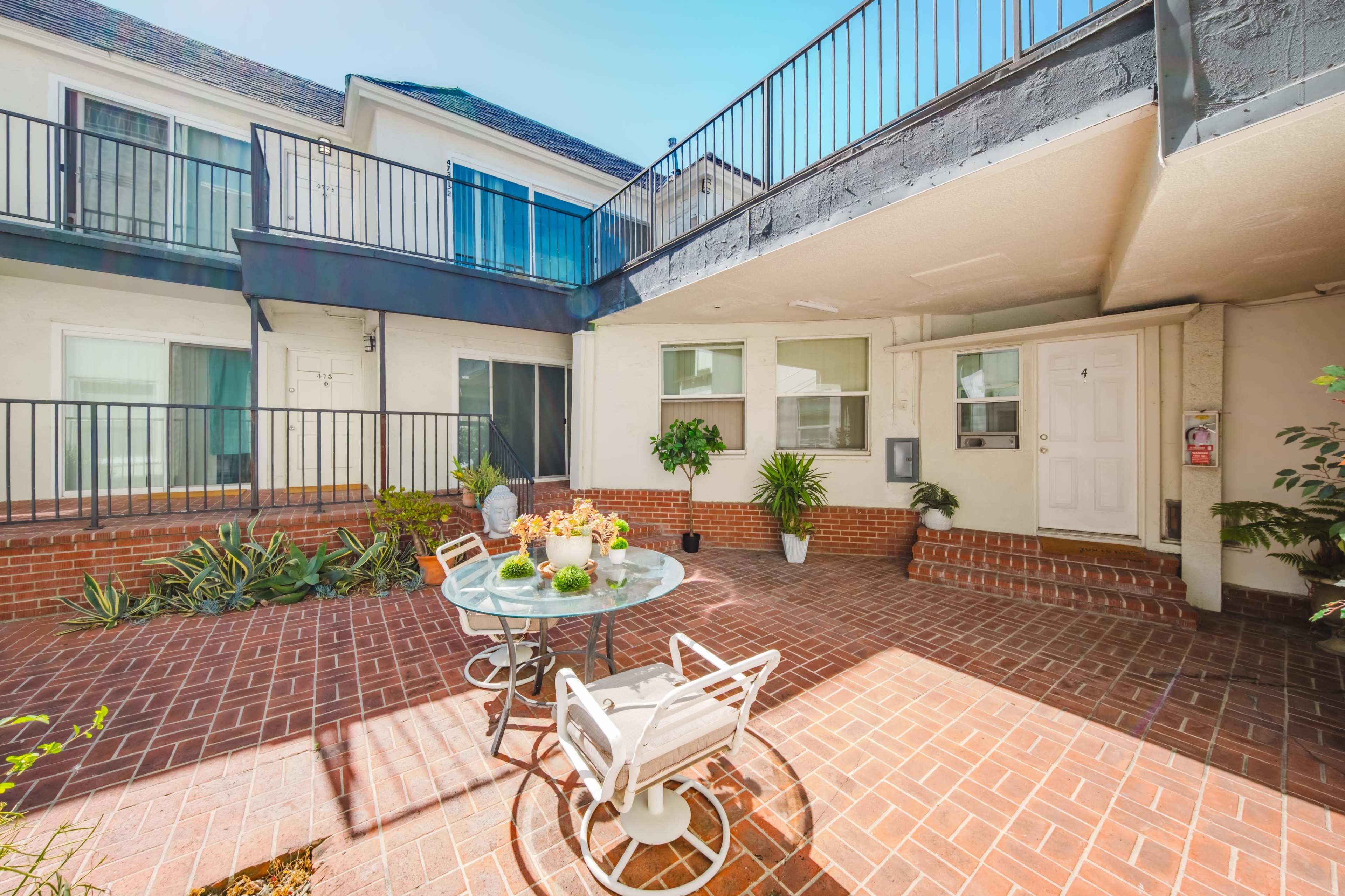 The image shows a small courtyard with a circular table and chairs surrounded by potted plants, adjacent to two-story buildings.