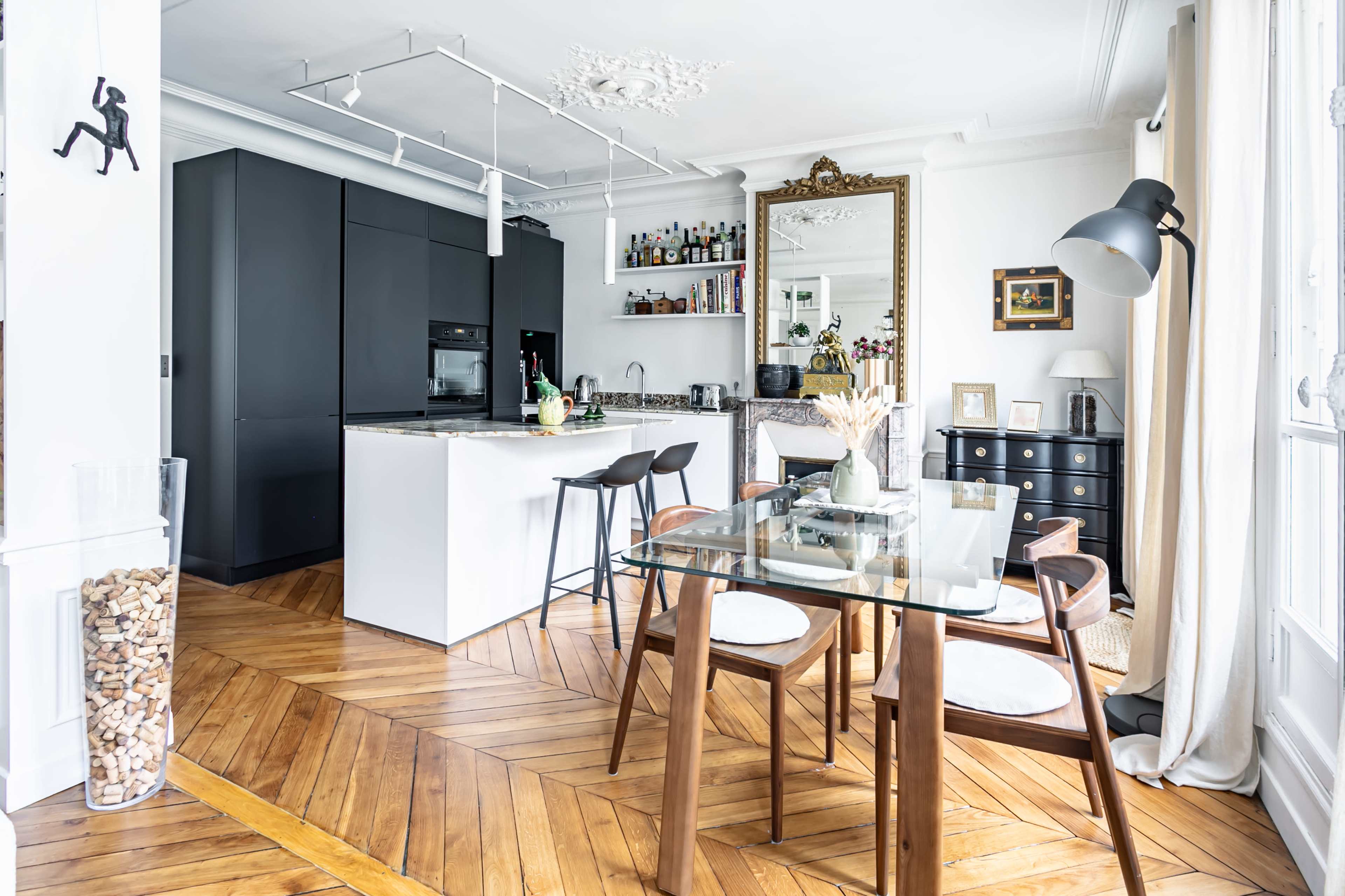 A modern kitchen and dining area features sleek black cabinetry, a glass-top dining table with wooden chairs, and a mirror reflecting the room's decor.