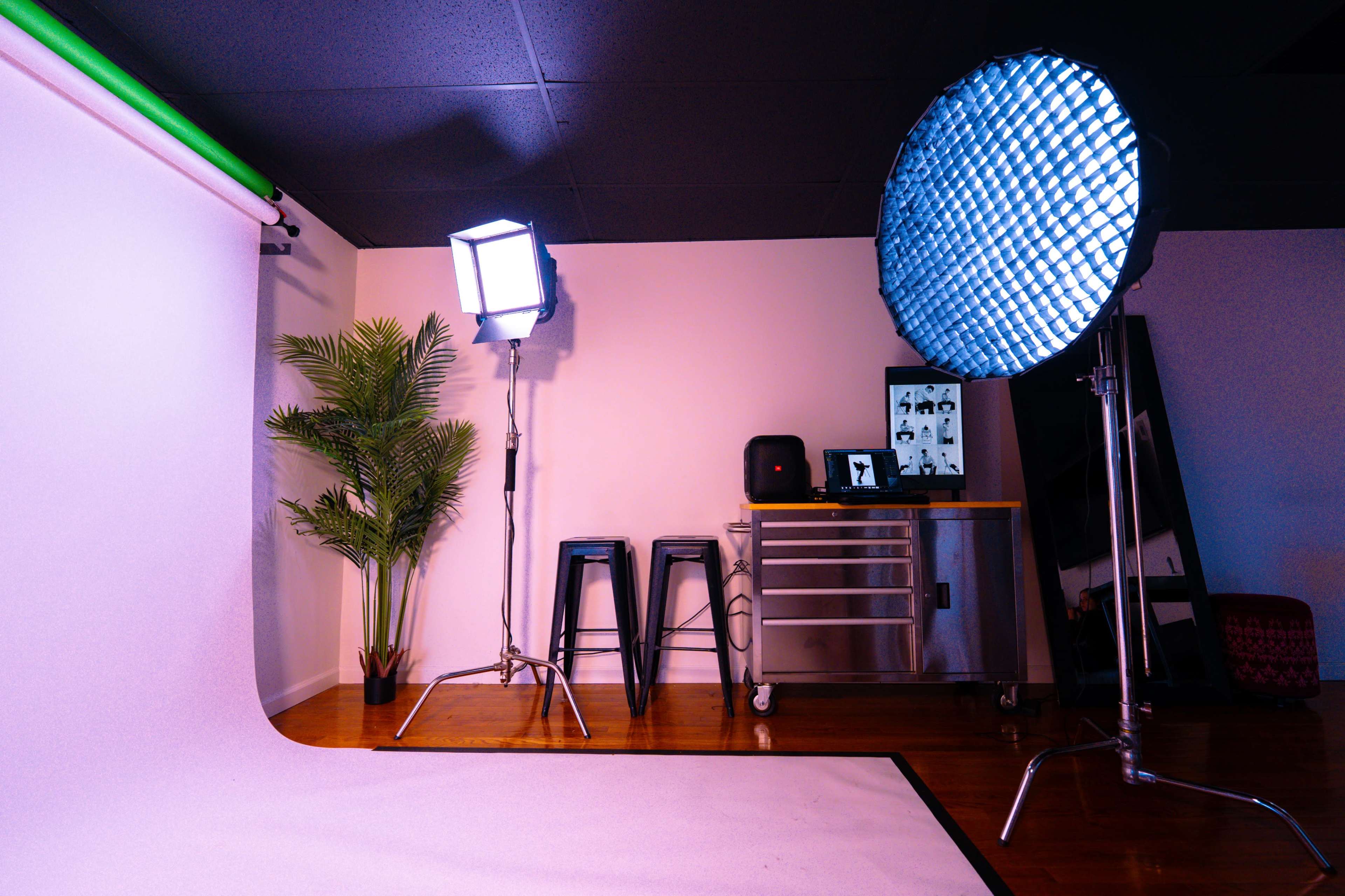 A photography studio features a green backdrop, lighting equipment, and a plant beside a table with a monitor displaying images.