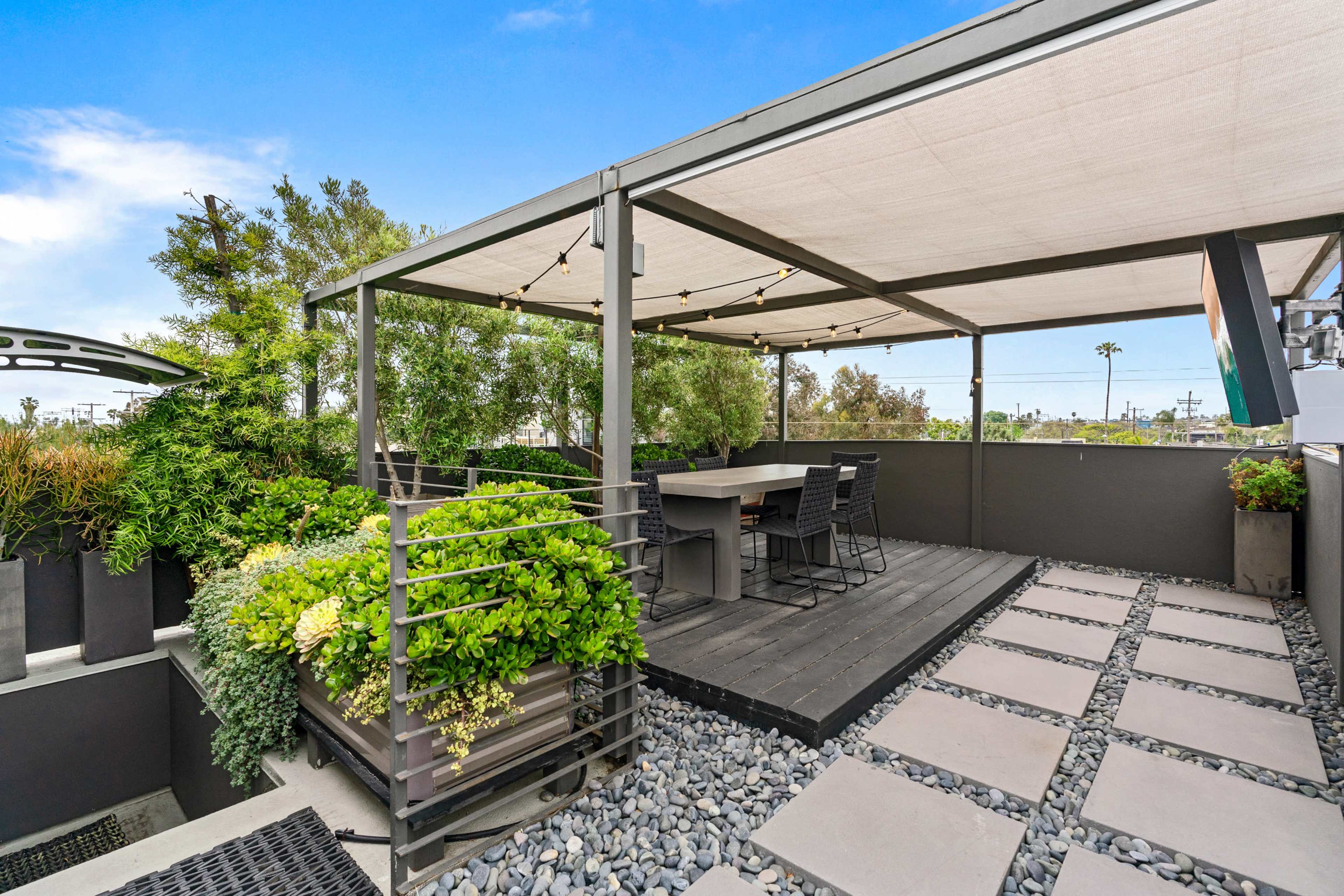 A modern rooftop patio with wooden decking, potted greenery, and a dining table surrounded by black chairs, all under a canopy with string lights.