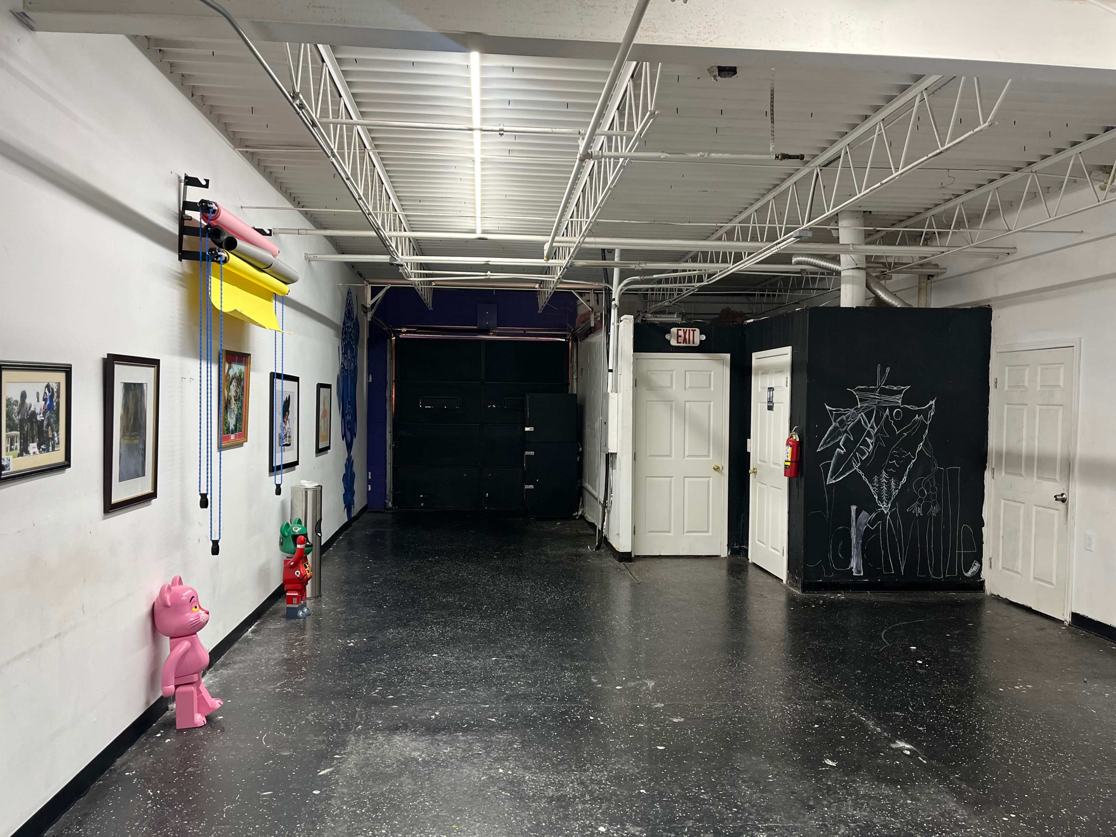 The image shows a spacious, well-lit interior with black flooring, white walls adorned with framed pictures, and a pink sculpture in the foreground.