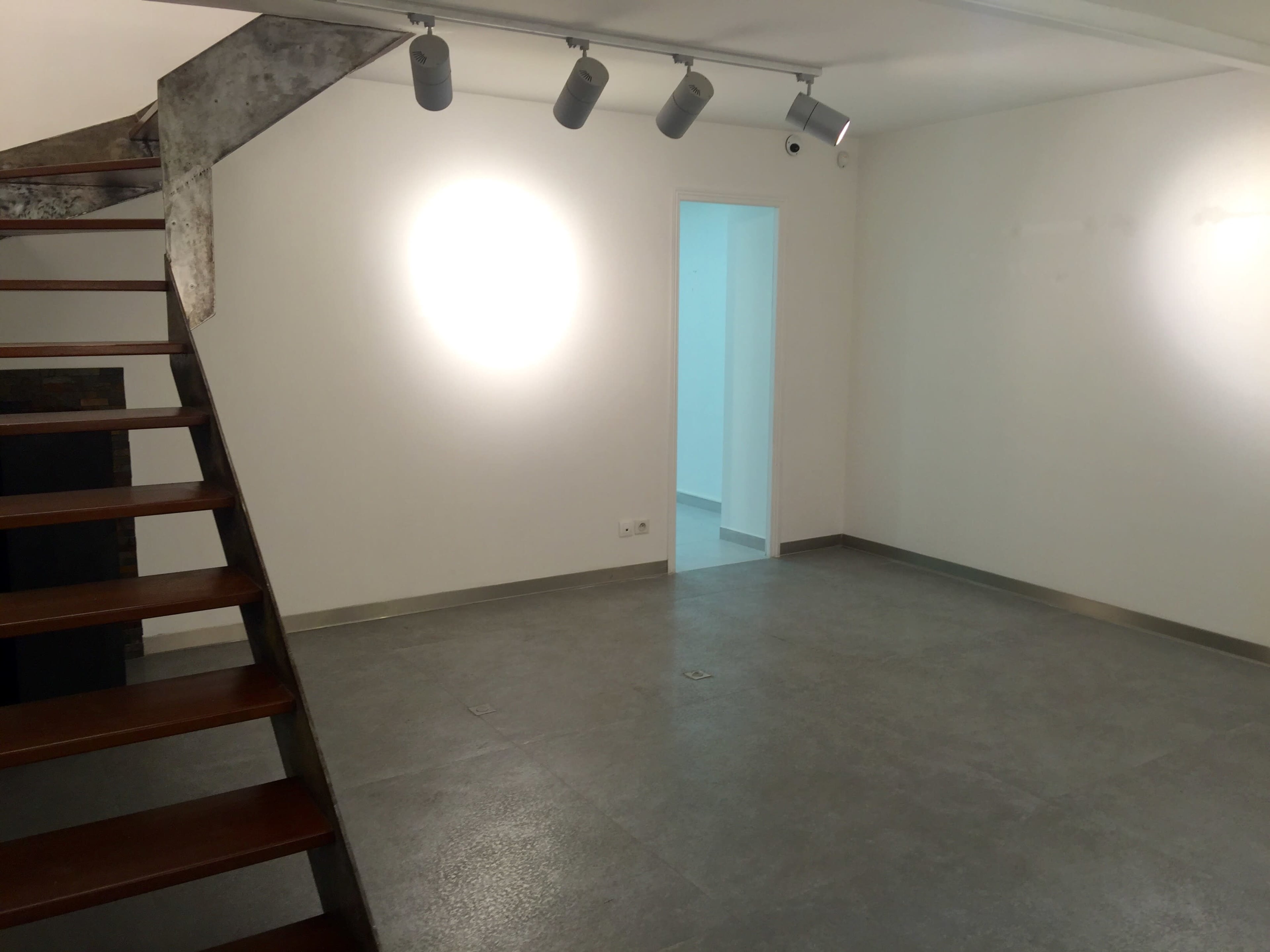 The image shows a minimalist interior space with a wooden staircase, concrete floor, and a doorway leading to another room.