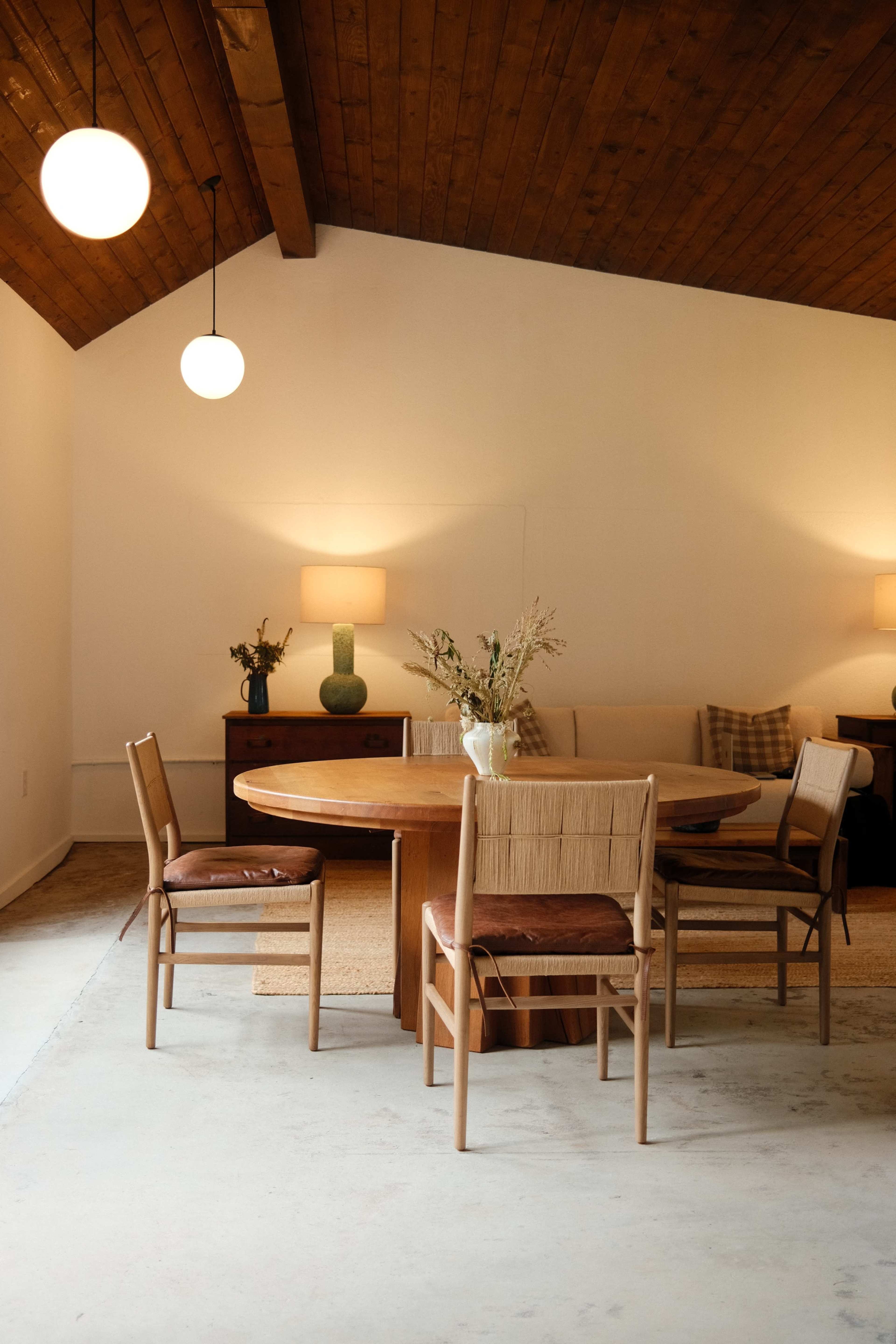 A wooden round dining table with four chairs is set in a room featuring a sloped wooden ceiling, soft lighting, and a neutral color scheme.