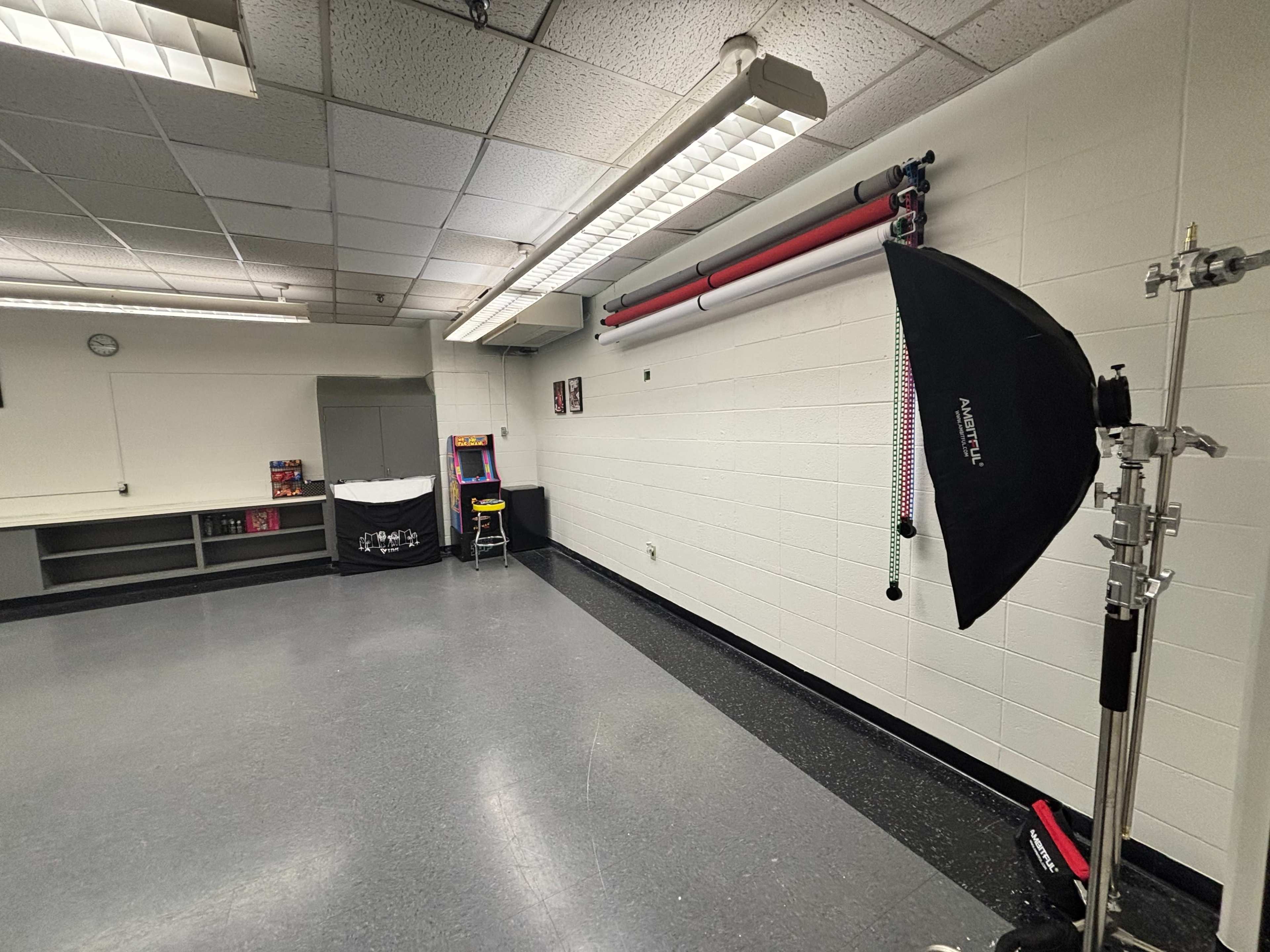 The image shows a spacious, well-lit room with gray floors and white walls, featuring a lighting setup, a small arcade machine, and a storage area.