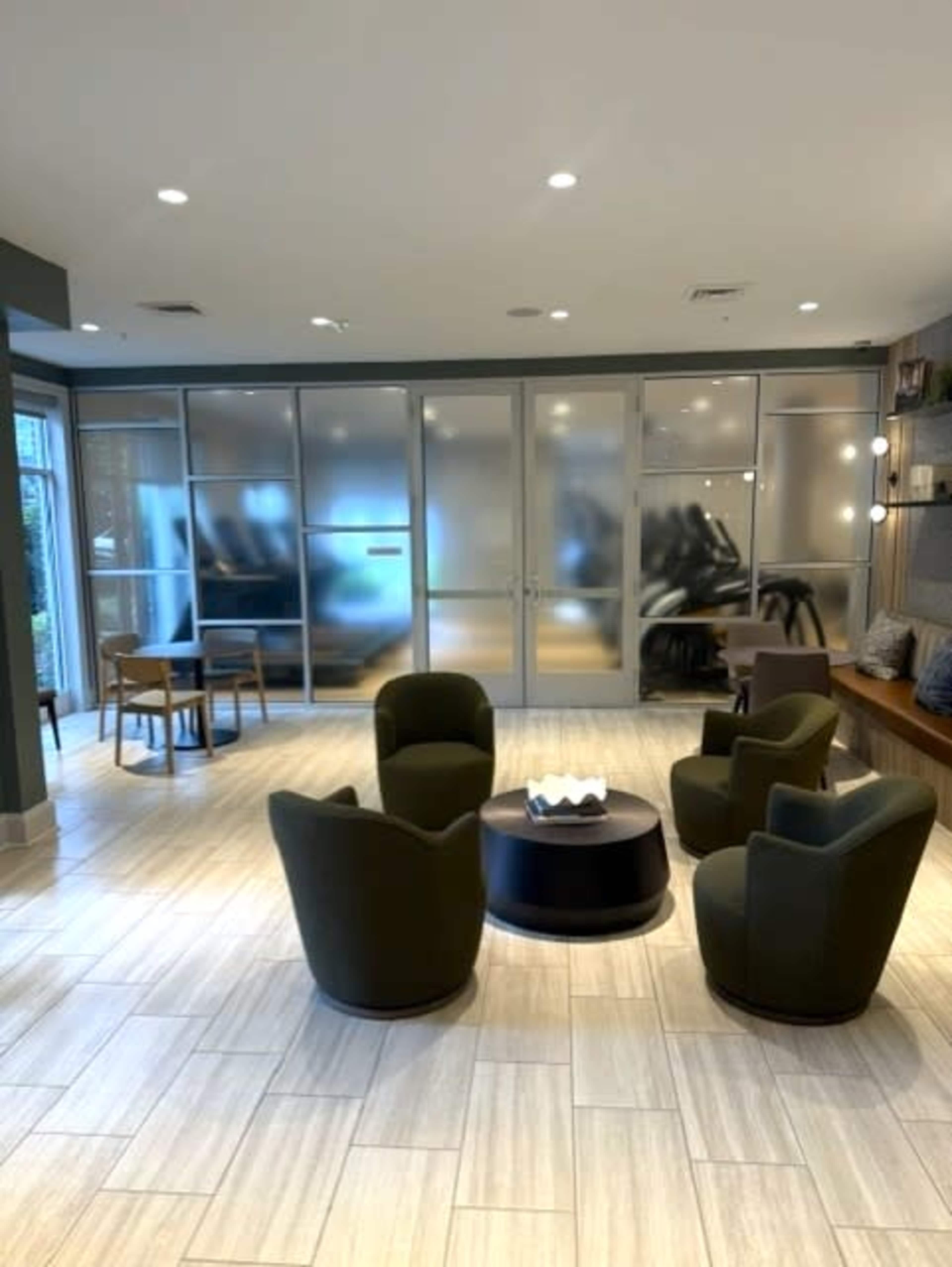 The image shows a modern lounge area featuring four green chairs arranged around a round table, with a glass partition in the background.