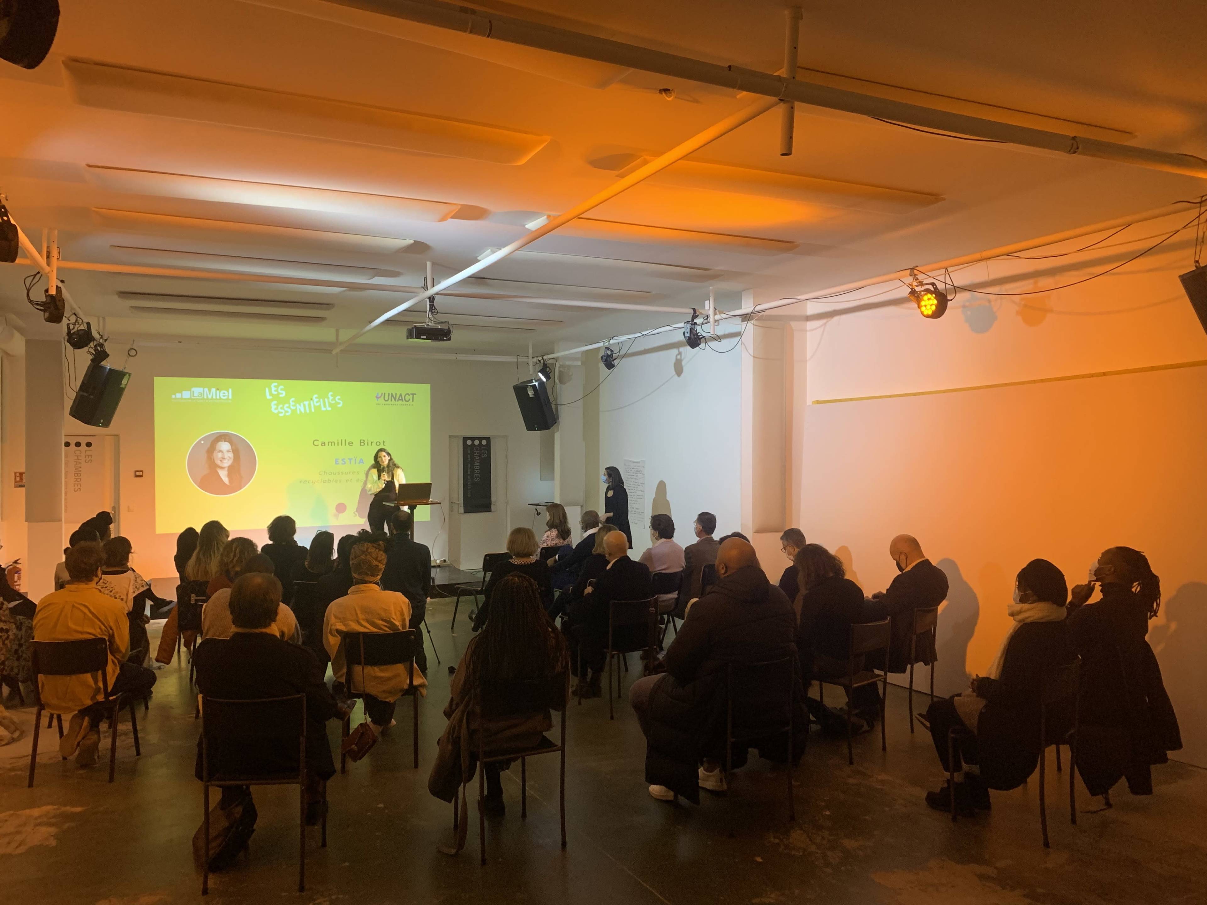 A speaker presents to an audience seated in a brightly lit room with a projector screen displaying information.