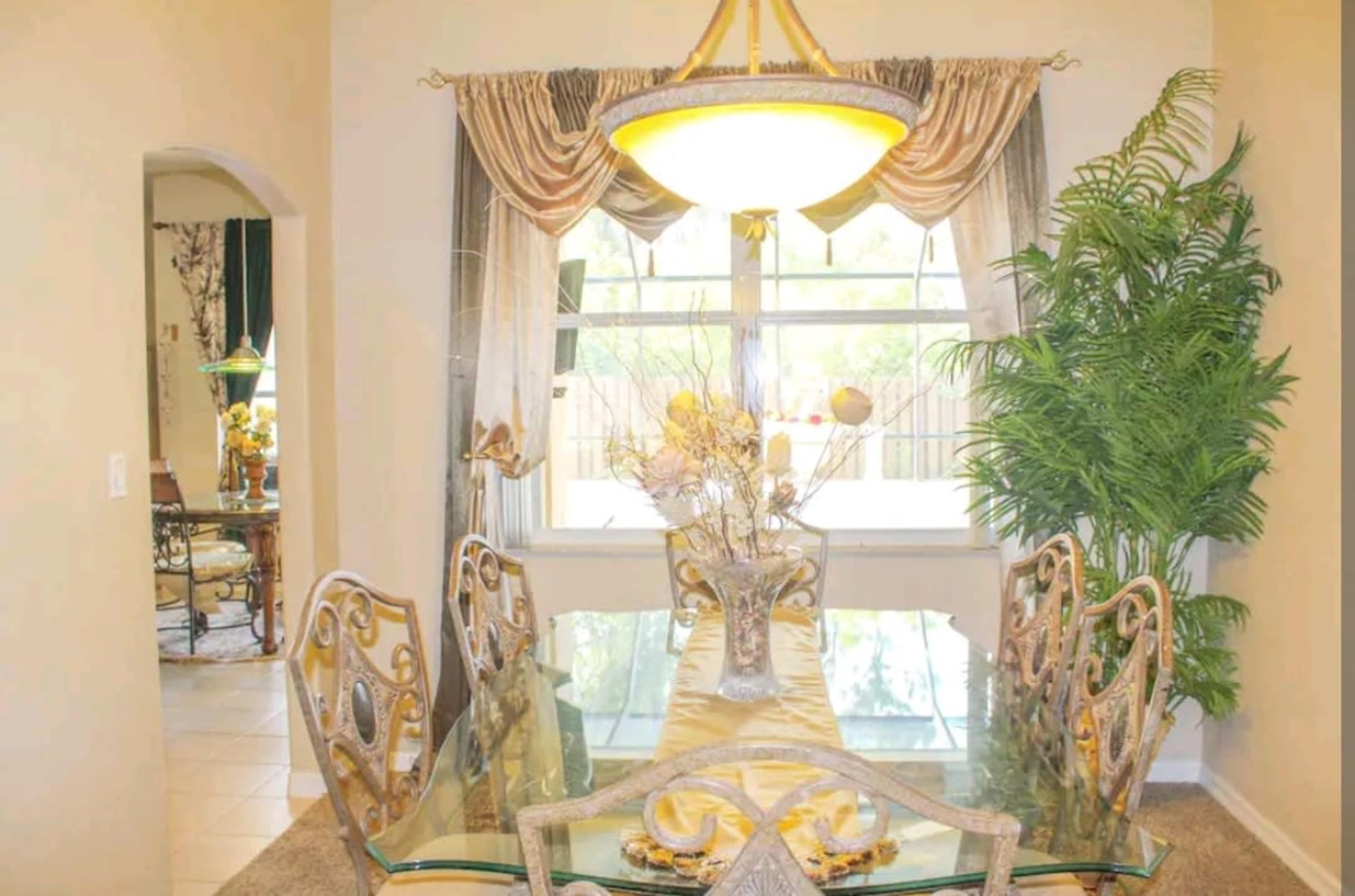 A dining room features a glass table surrounded by six ornate chairs, with a large light fixture above and a potted plant in the corner.