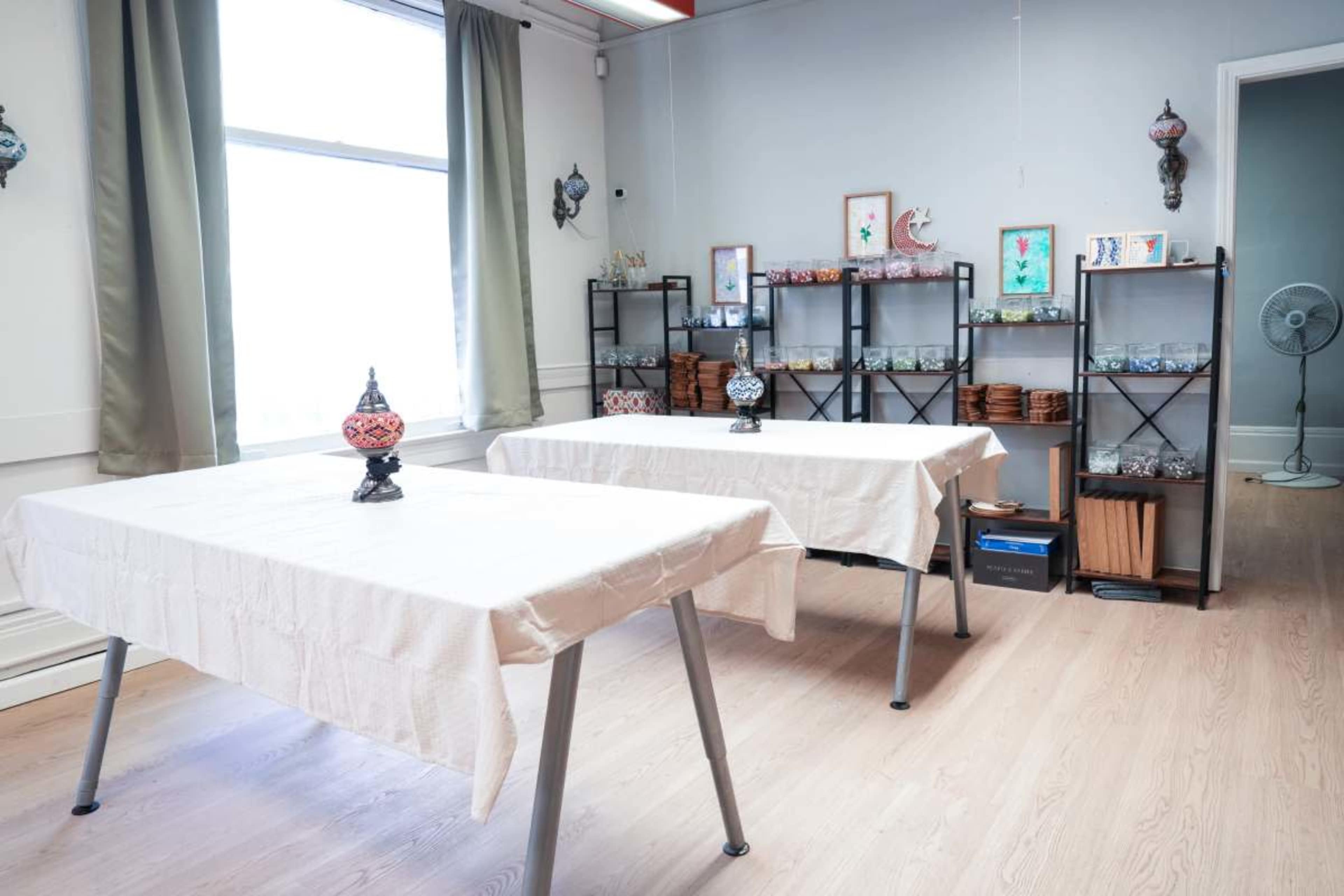 The image shows a bright, uncluttered room with two tables covered in light-colored tablecloths and shelves displaying decorative items along the walls.