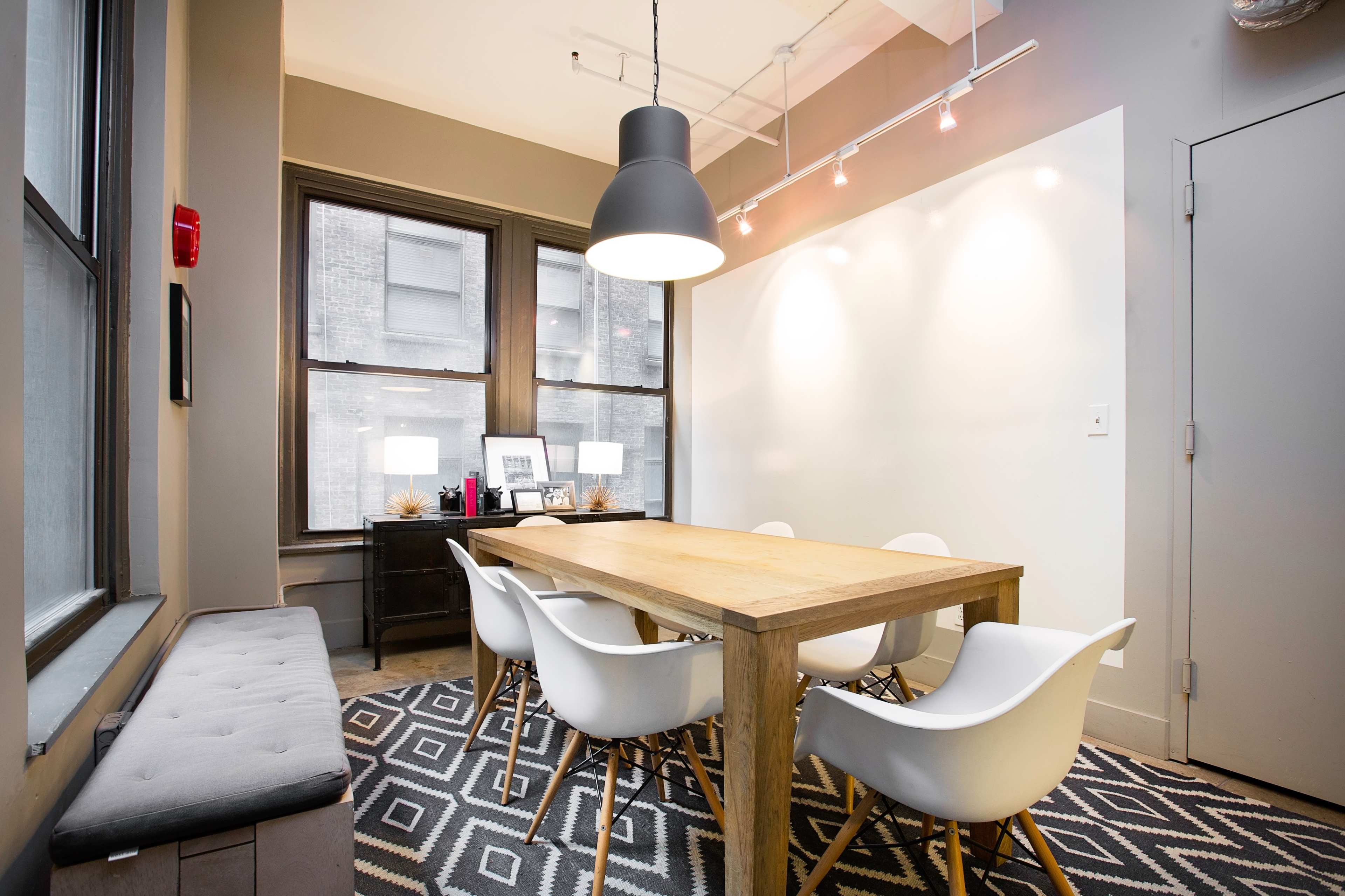 A meeting room features a large wooden table surrounded by white chairs, with windows showcasing a city view and a geometric-patterned rug on the floor.