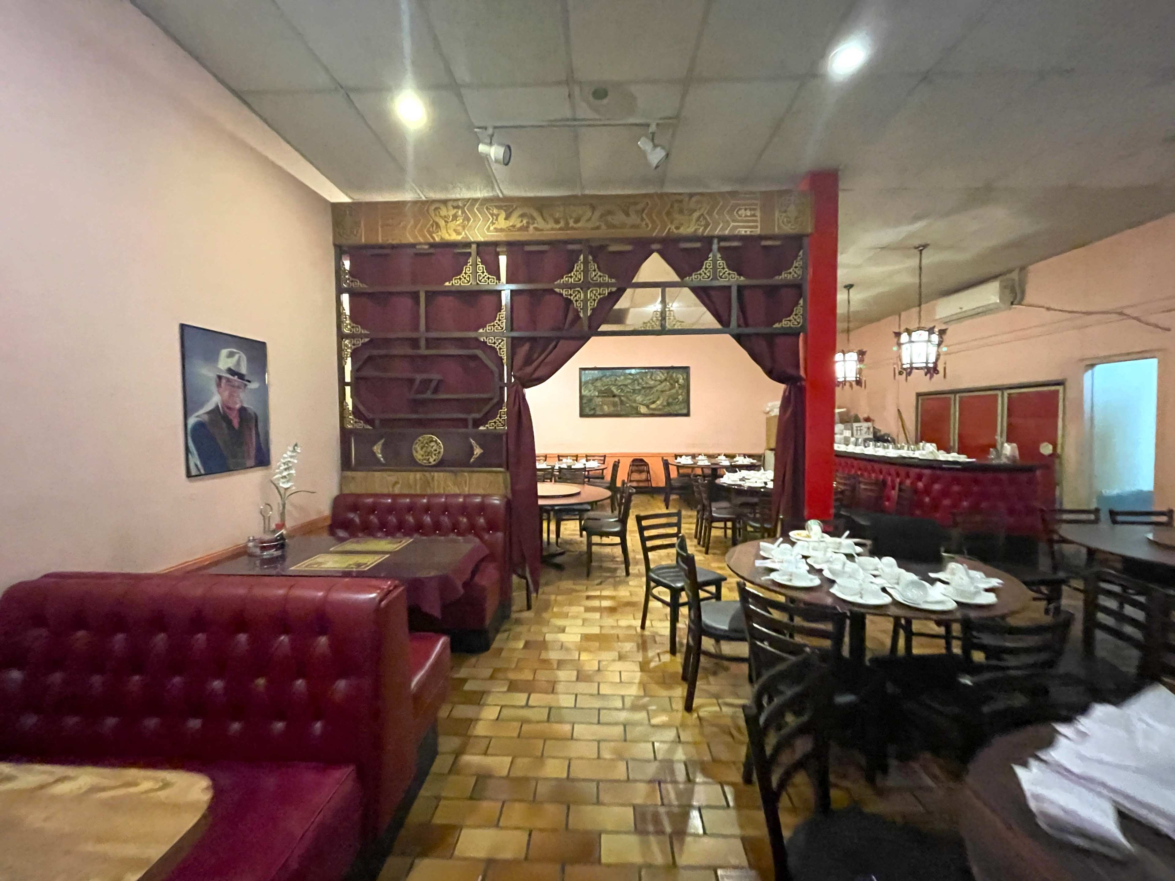 The image shows an empty restaurant interior with red booths, wooden tables, and a decorative divider, featuring a painting on the wall and white table settings.