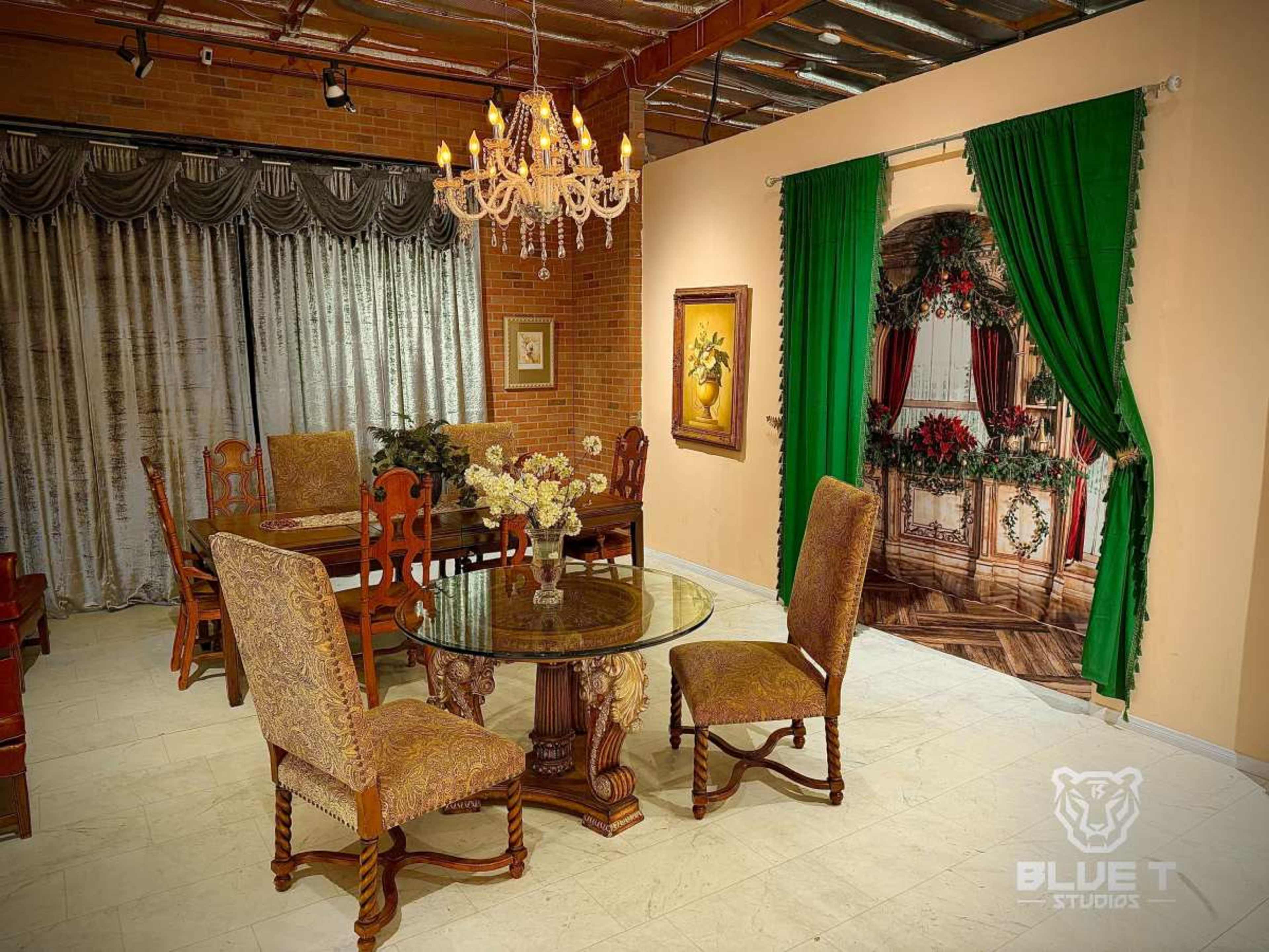 The image features an elegantly arranged dining area with a round glass table, ornate chairs, and decorative curtains, complemented by a chandelier overhead and an artistic backdrop of a room adorned for the holidays.
