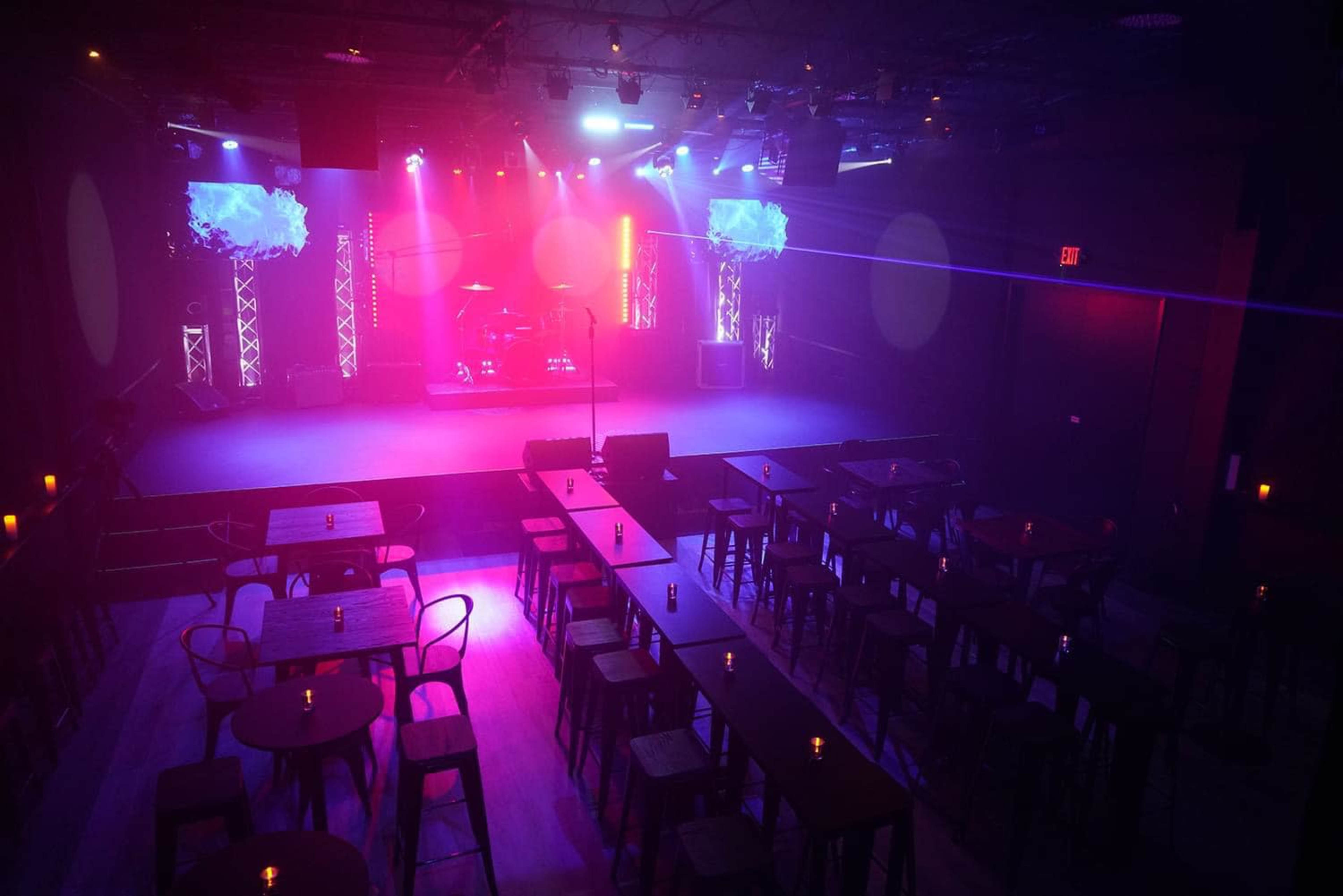 A dimly lit performance space with rows of tables and chairs facing a stage illuminated by colorful lights.